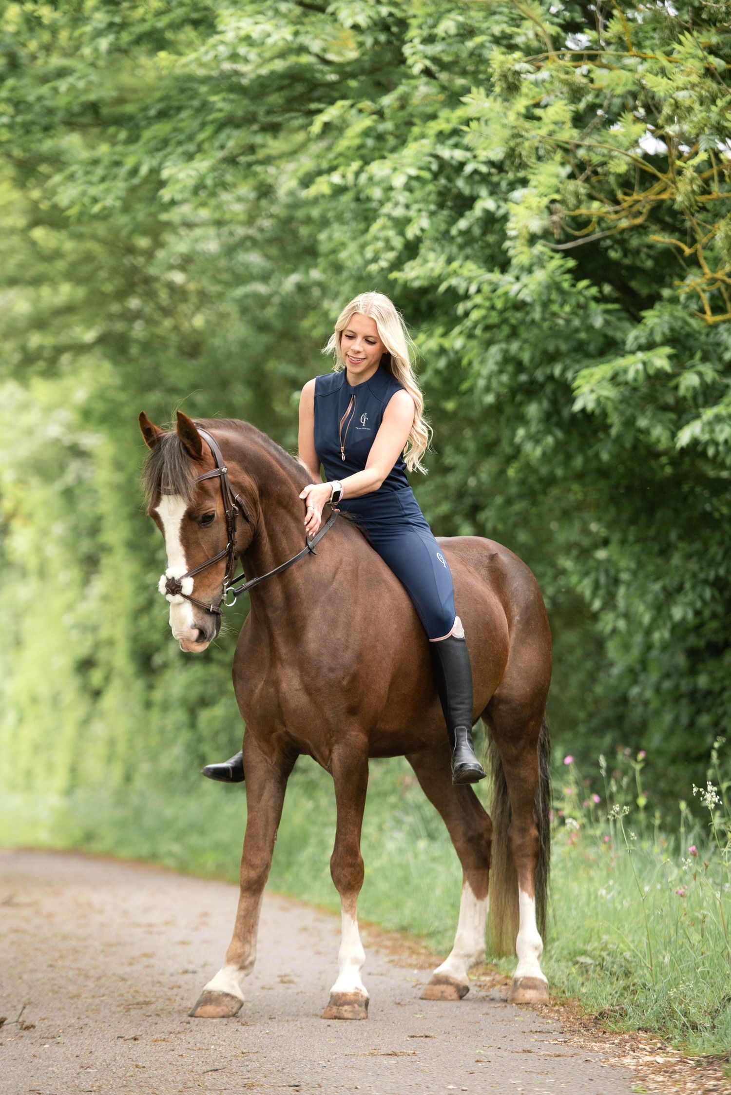 Blonde equestrian in navy riding outfit atop a chestnut horse on a scenic green country lane.