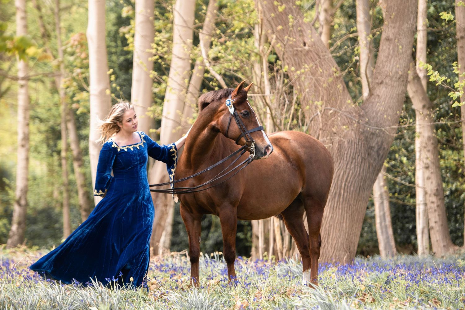 Woman in blue medieval gown leading a chestnut horse through a bluebell woodland forest.