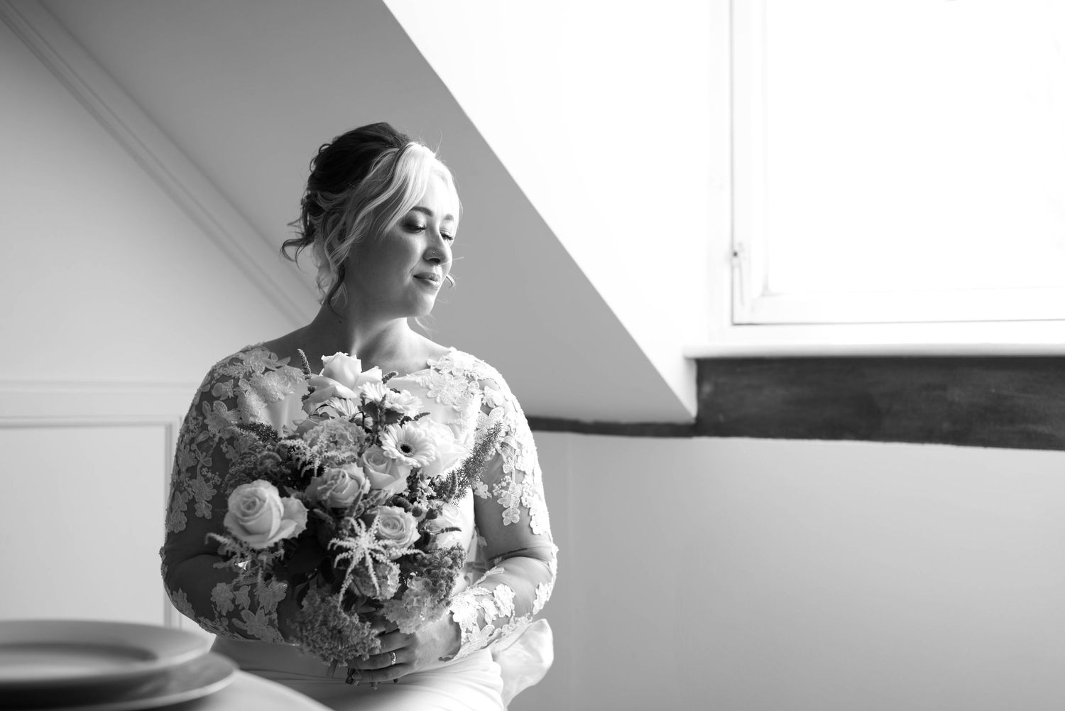 Black and white portrait of bride in lace wedding dress holding bouquet near window in soft natural light.