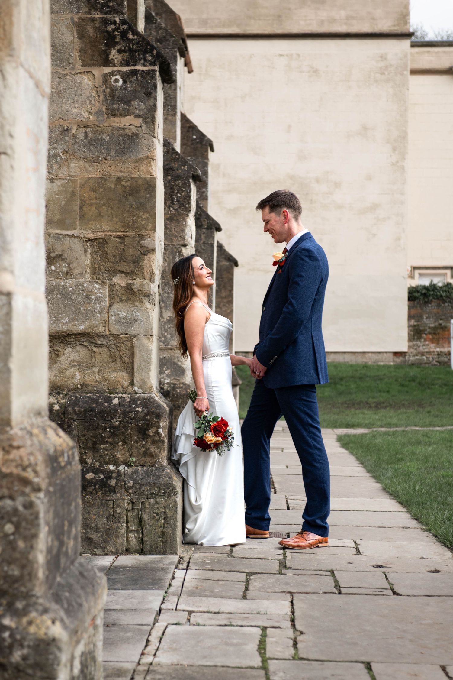 Bride in white gown and groom in navy suit hold hands beside stone church columns.
