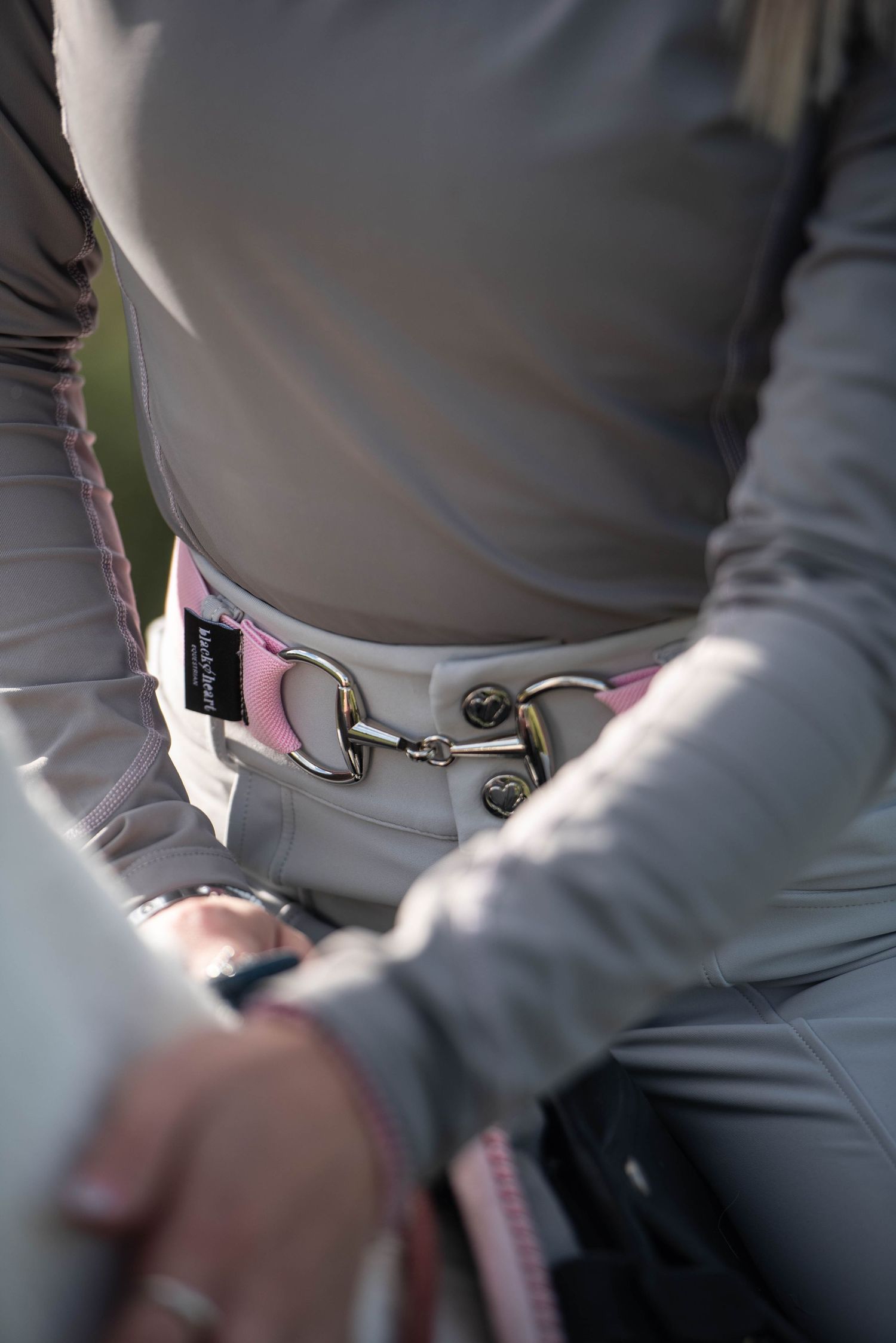 Close-up of an equestrian rider wearing a grey top and white breeches with a decorative pink snaffle bit belt.