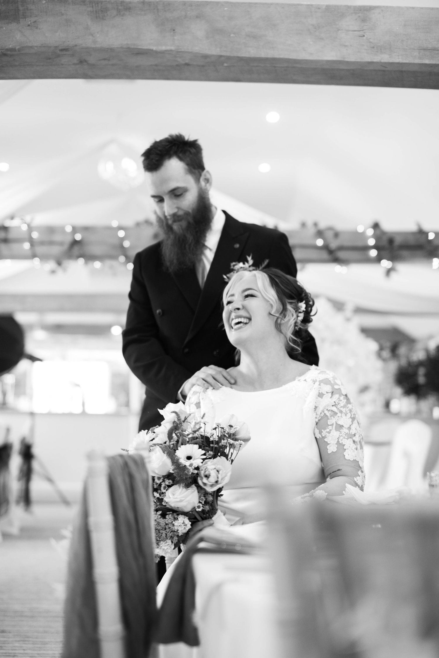 A black and white photo of a laughing bride holding flowers with her bearded groom at their wedding.