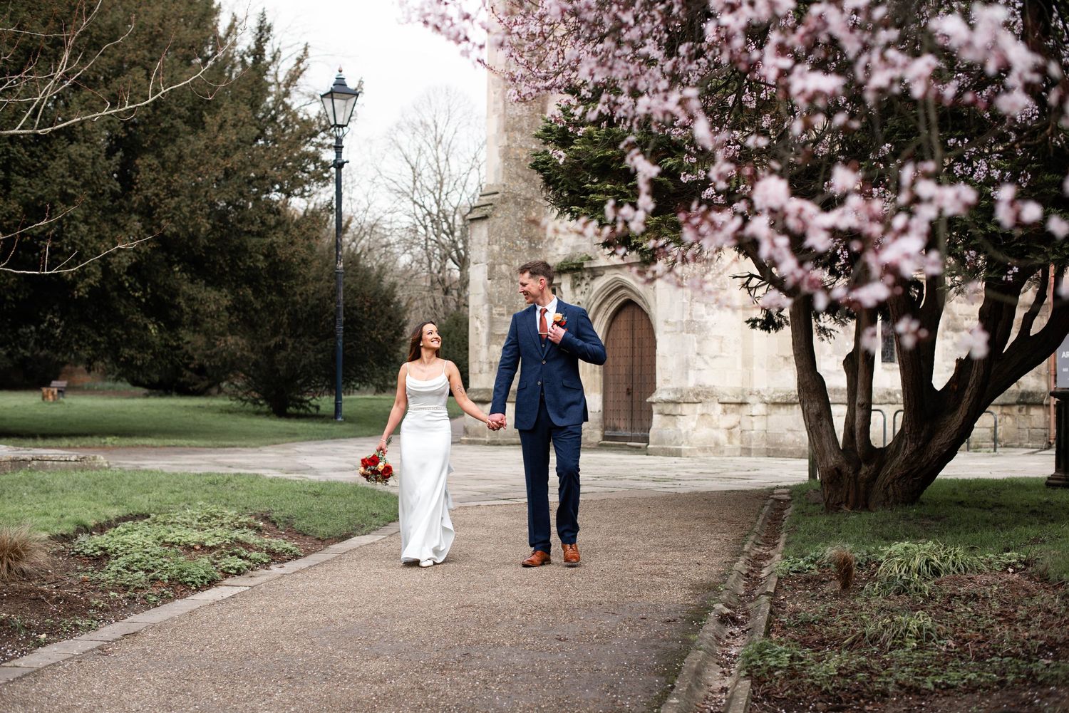 Bride and groom walking hand in hand on a garden path beside a blooming cherry blossom tree near a historic stone chapel.