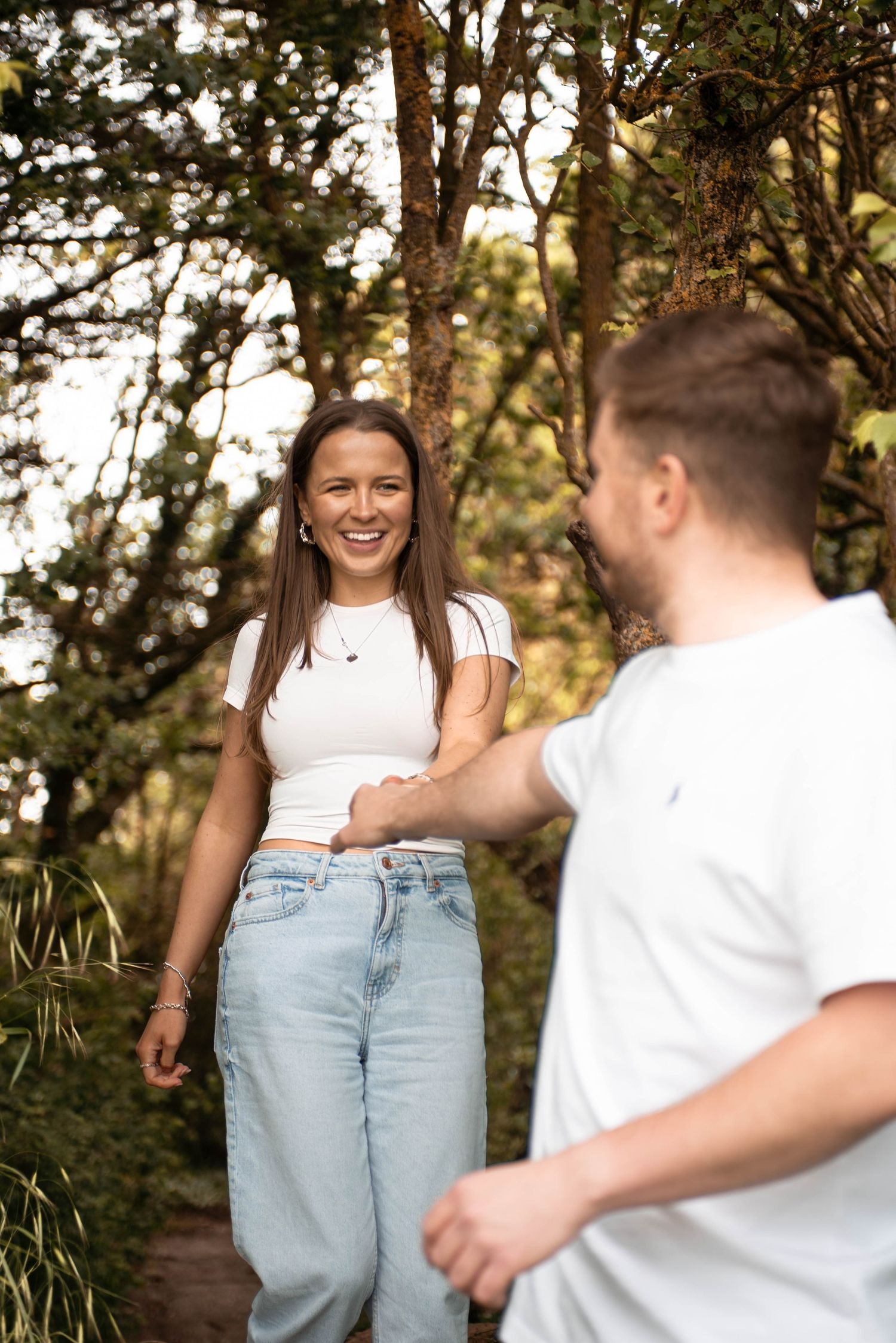 Happy couple holding hands on a forest path, both wearing white tops and jeans.