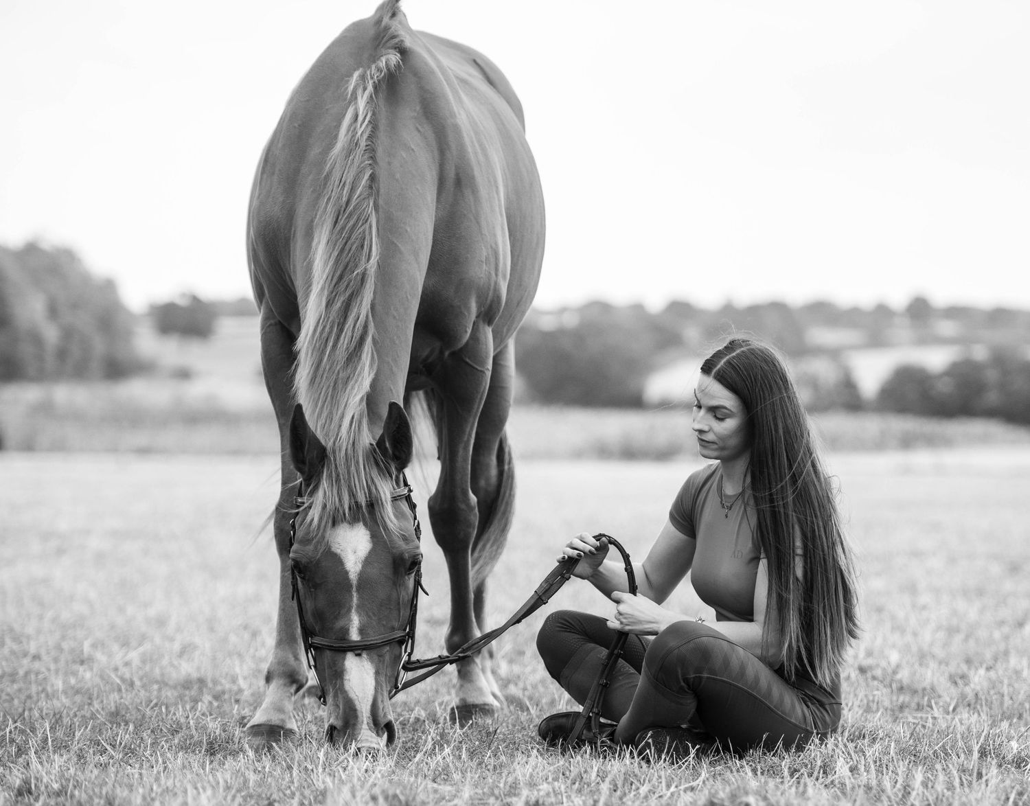 Black and white photo of a woman sitting on grass beside a grazing horse in an open field.