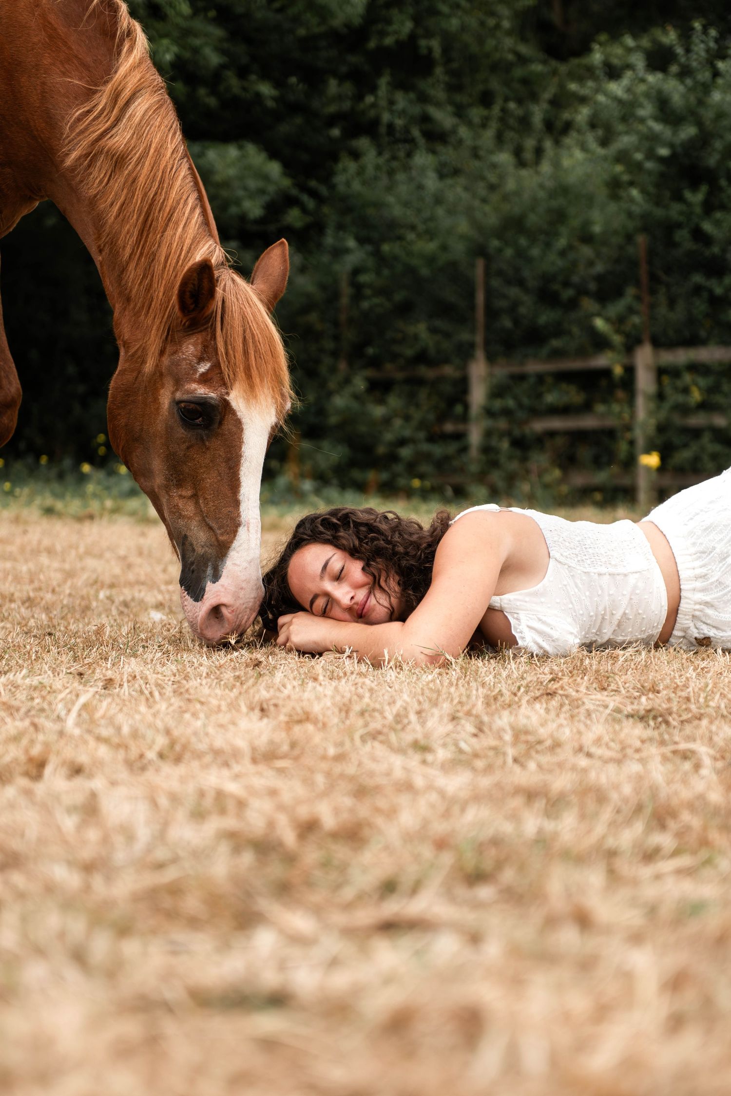Young woman in white dress lying on dry grass, smiling with eyes closed beside a chestnut horse grazing in a field.