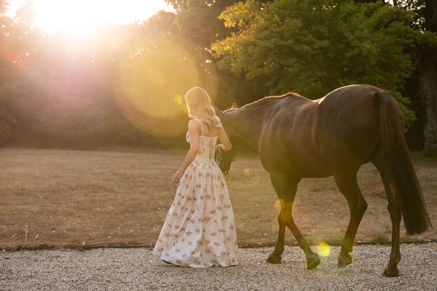 A woman in a floral gown walks beside a brown horse at golden hour in a sunlit outdoor setting.