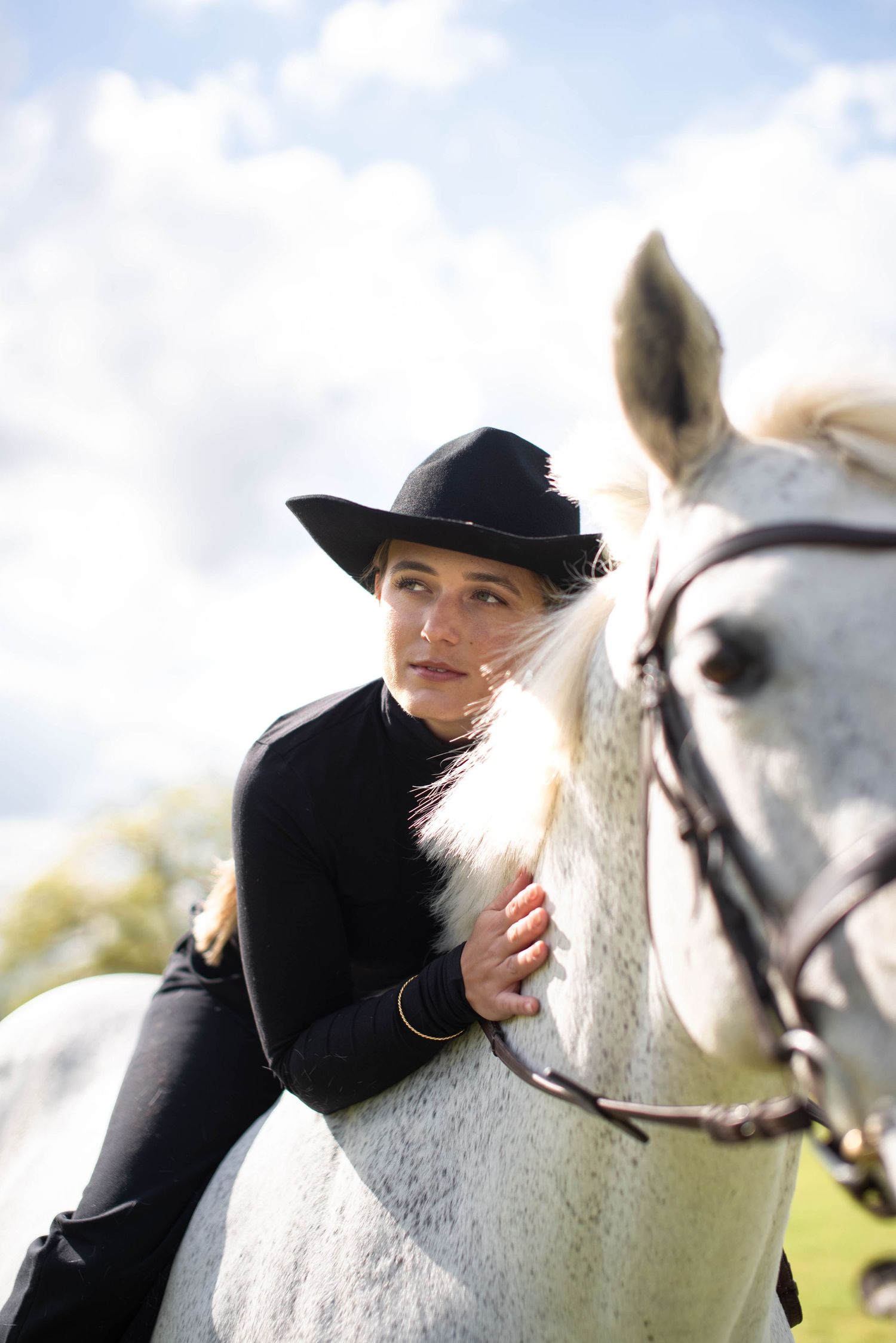 Woman in black outfit and cowboy hat leans affectionately on a white horse outdoors.