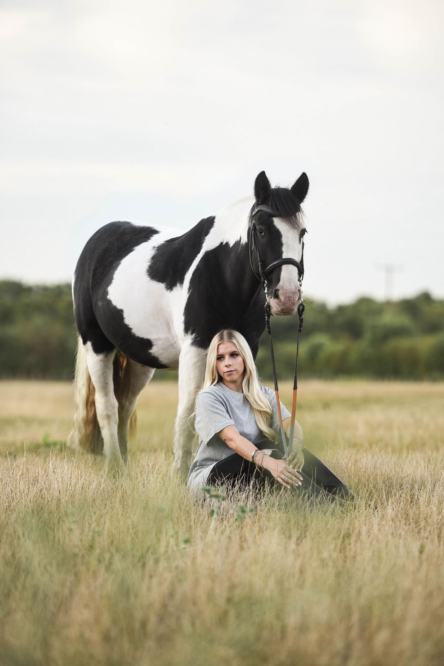 Blonde woman sitting in golden field beside tall black and white paint horse.