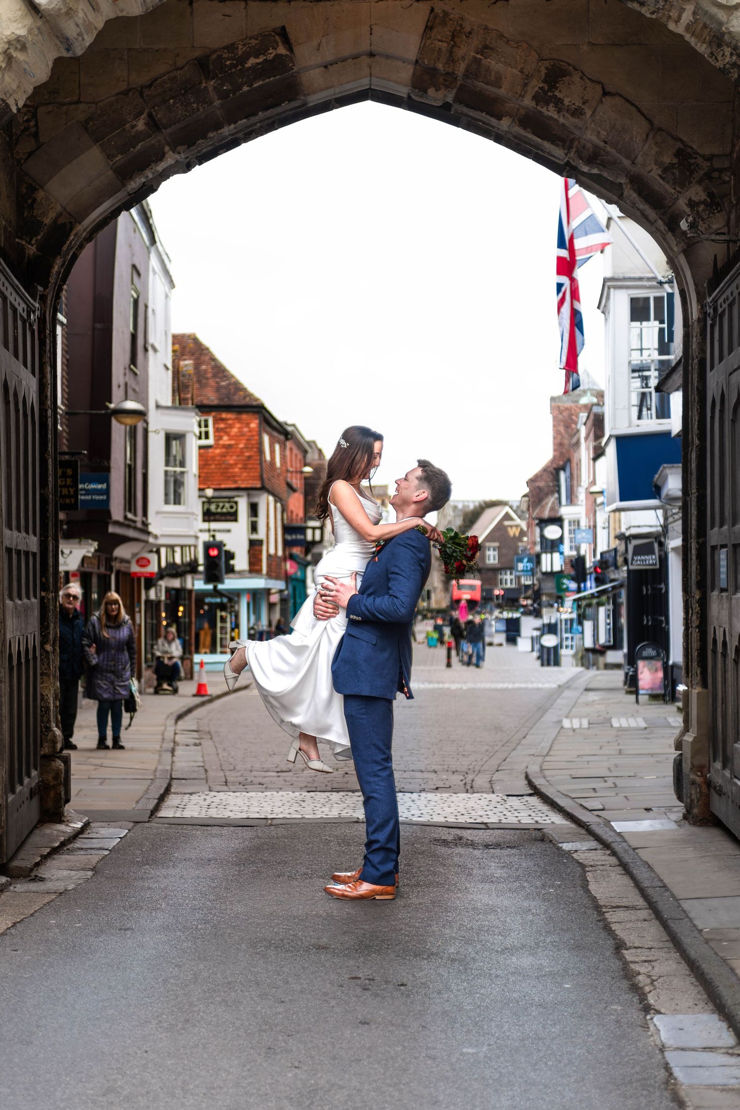 Groom lifts bride in wedding dress under historic arch gateway on a UK high street.