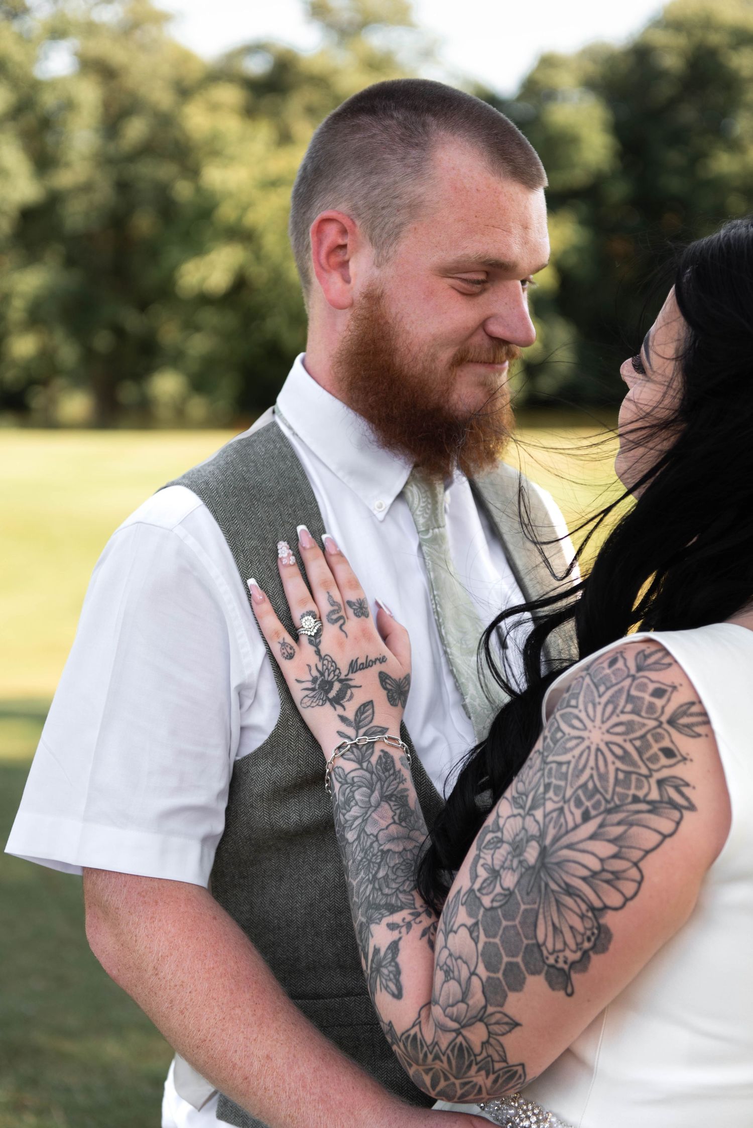 A tattooed couple shares a tender moment outdoors, the man in a grey vest and tie, the woman with sleeve tattoos embracing him.