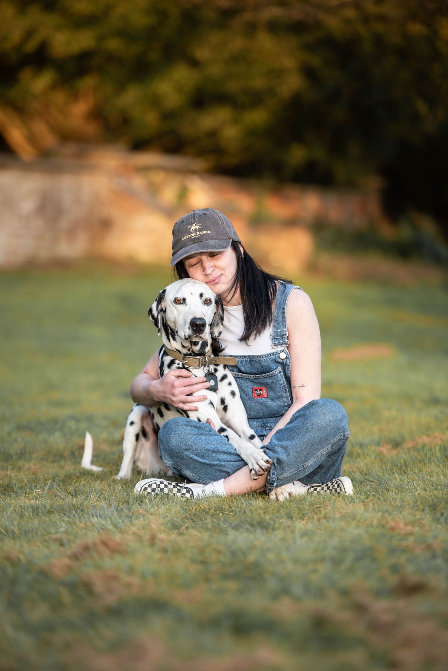 A young woman in overalls and cap hugging her Dalmatian dog on green grass.