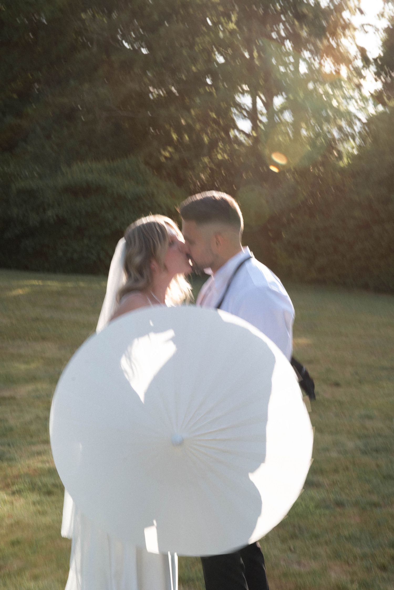 Bride and groom share a romantic moment under golden sunset light, holding a white parasol outdoors.