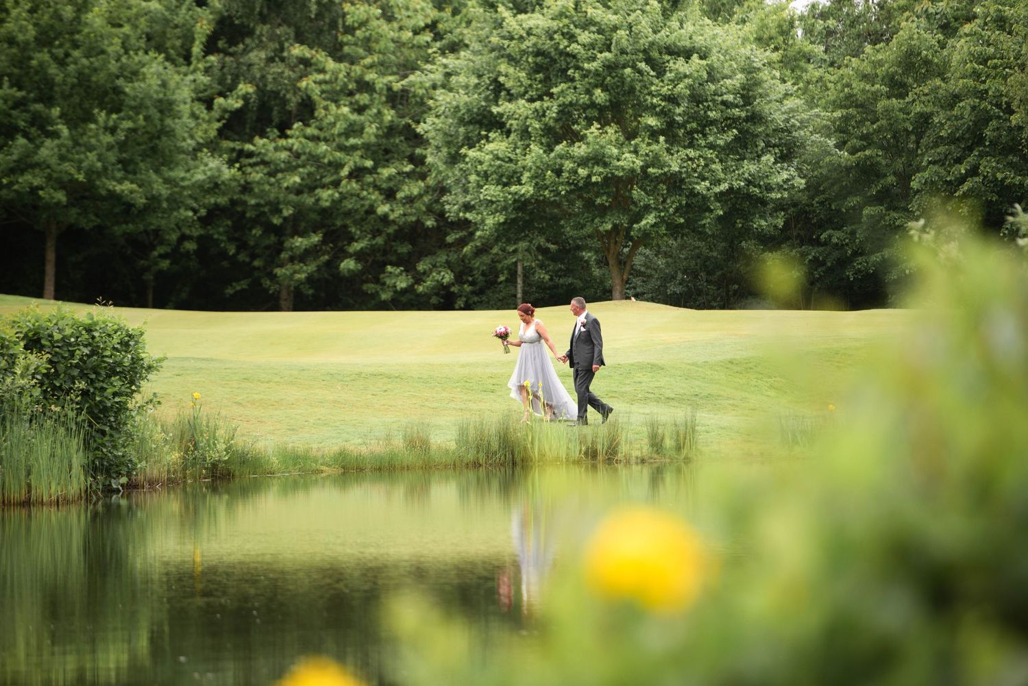 A bride and groom walk hand in hand along a serene pond surrounded by lush green trees.