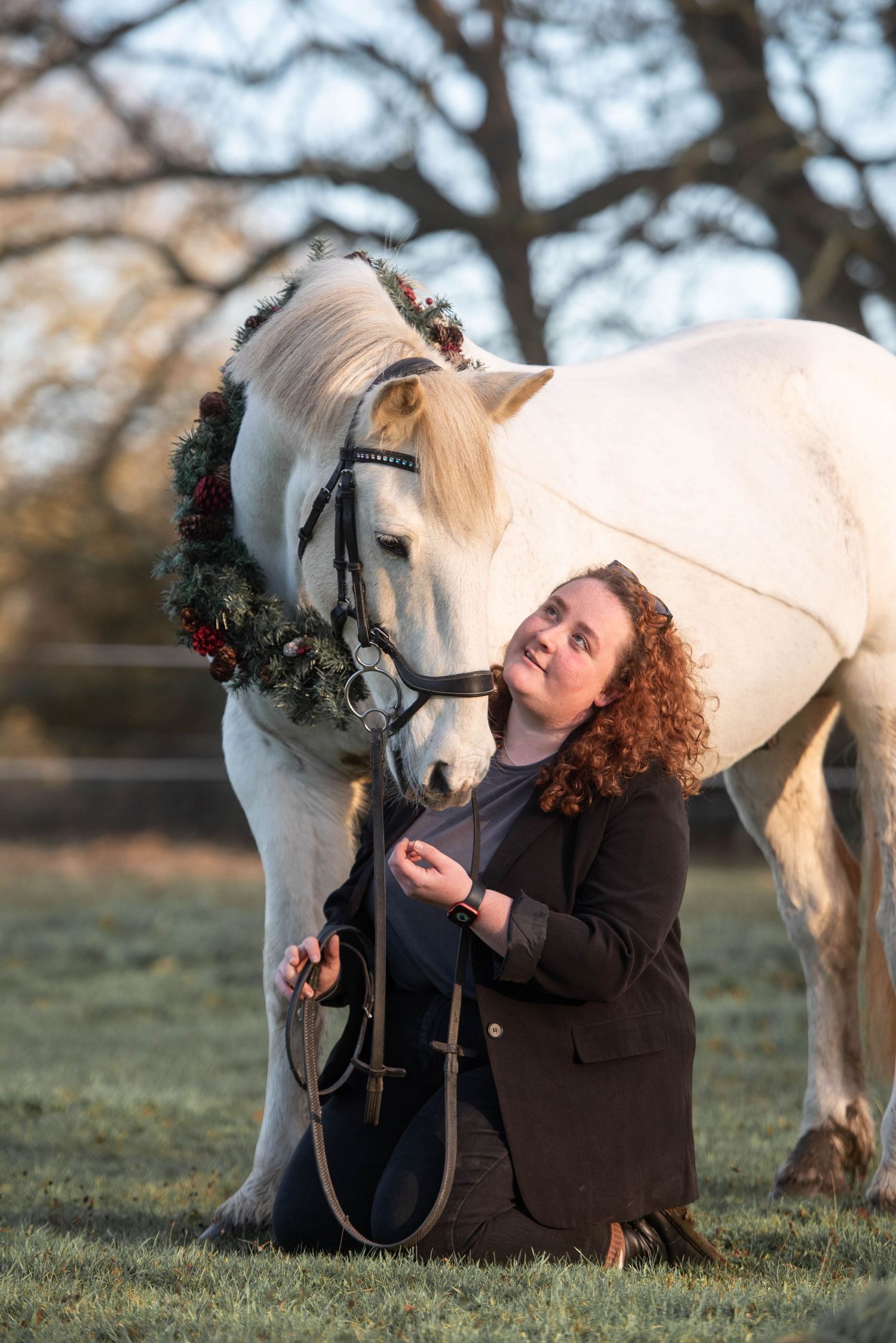 Woman with curly hair kneeling beside a white horse wearing a festive holiday wreath in a field.