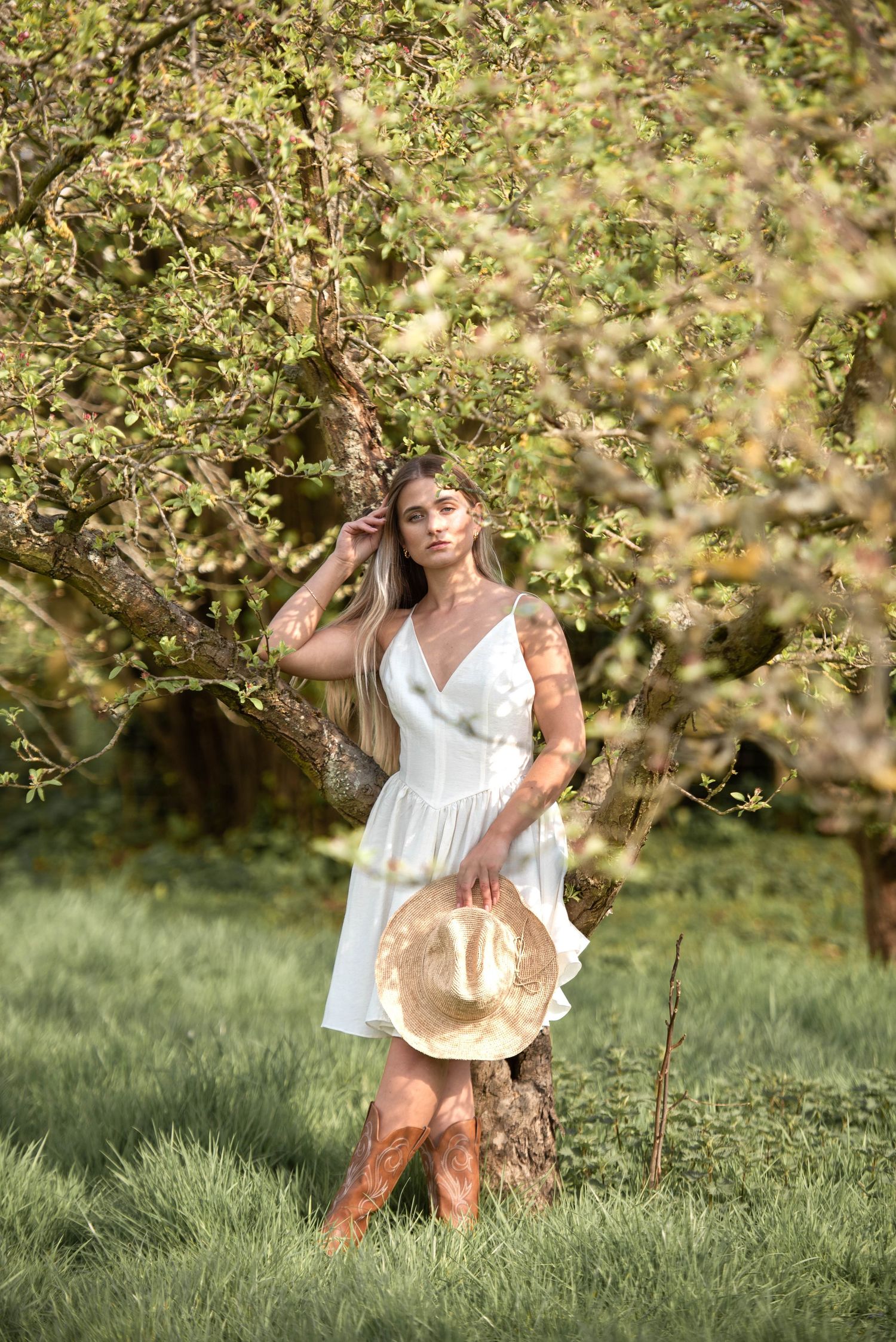 Woman in white dress and cowboy boots holds straw hat near blossoming tree in golden sunlight.