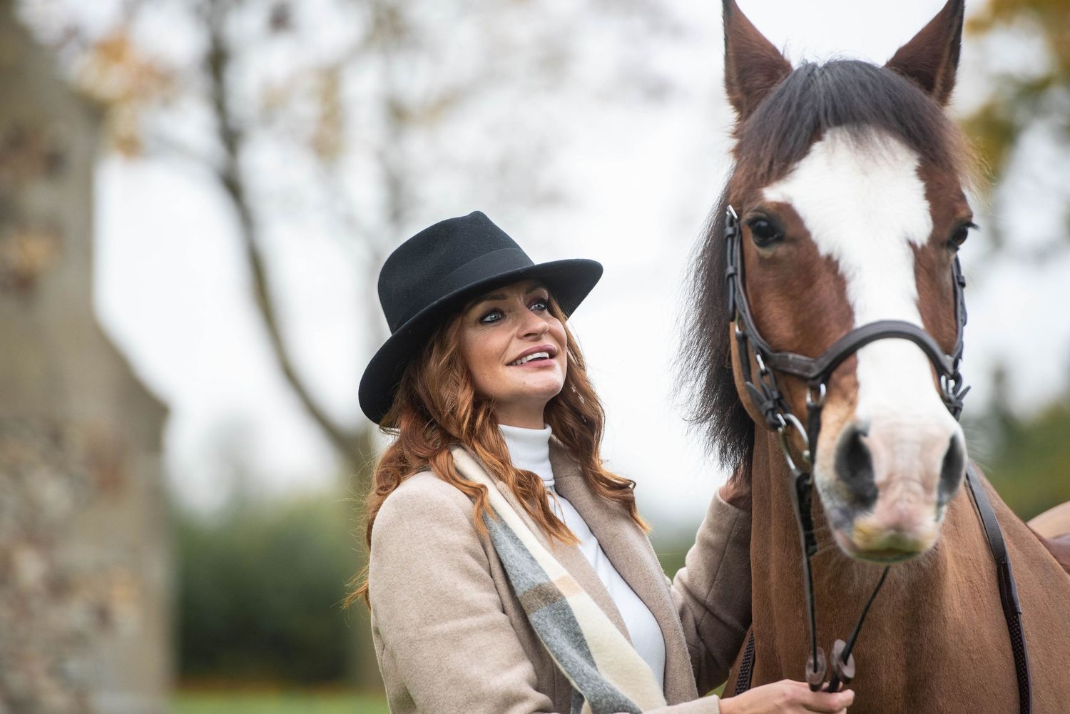 Smiling woman in a black hat standing beside a chestnut horse with a white blaze outdoors.