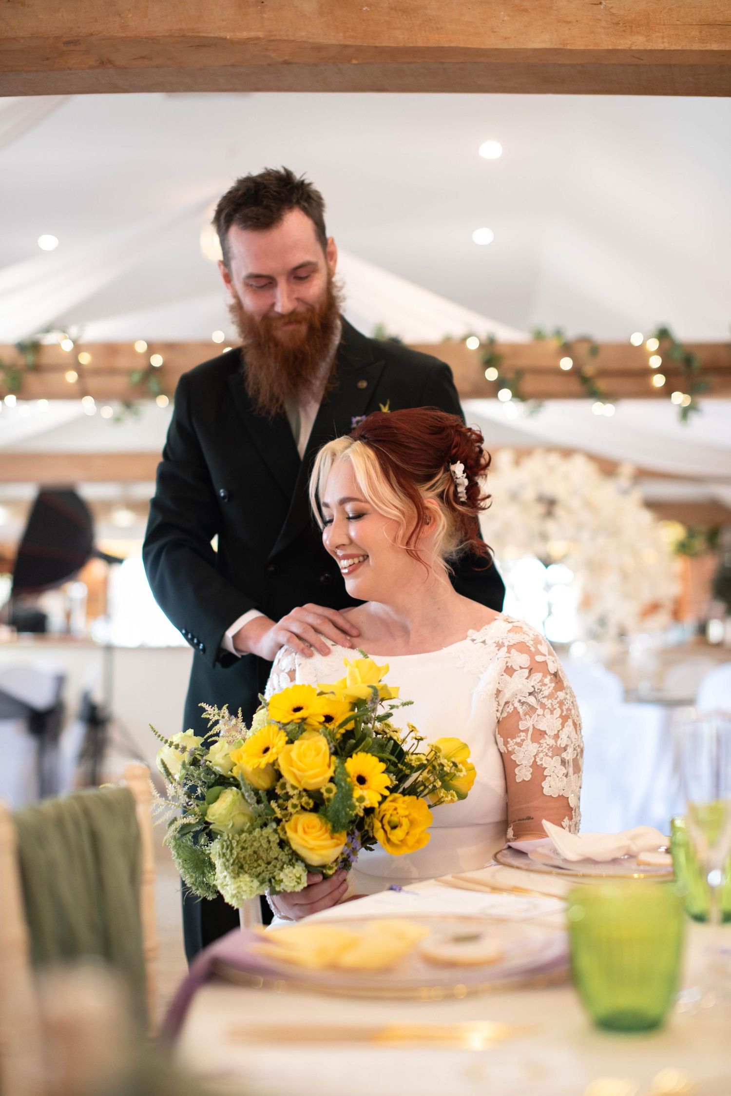 A bearded groom places his hand on a smiling bride holding a yellow floral bouquet at reception.
