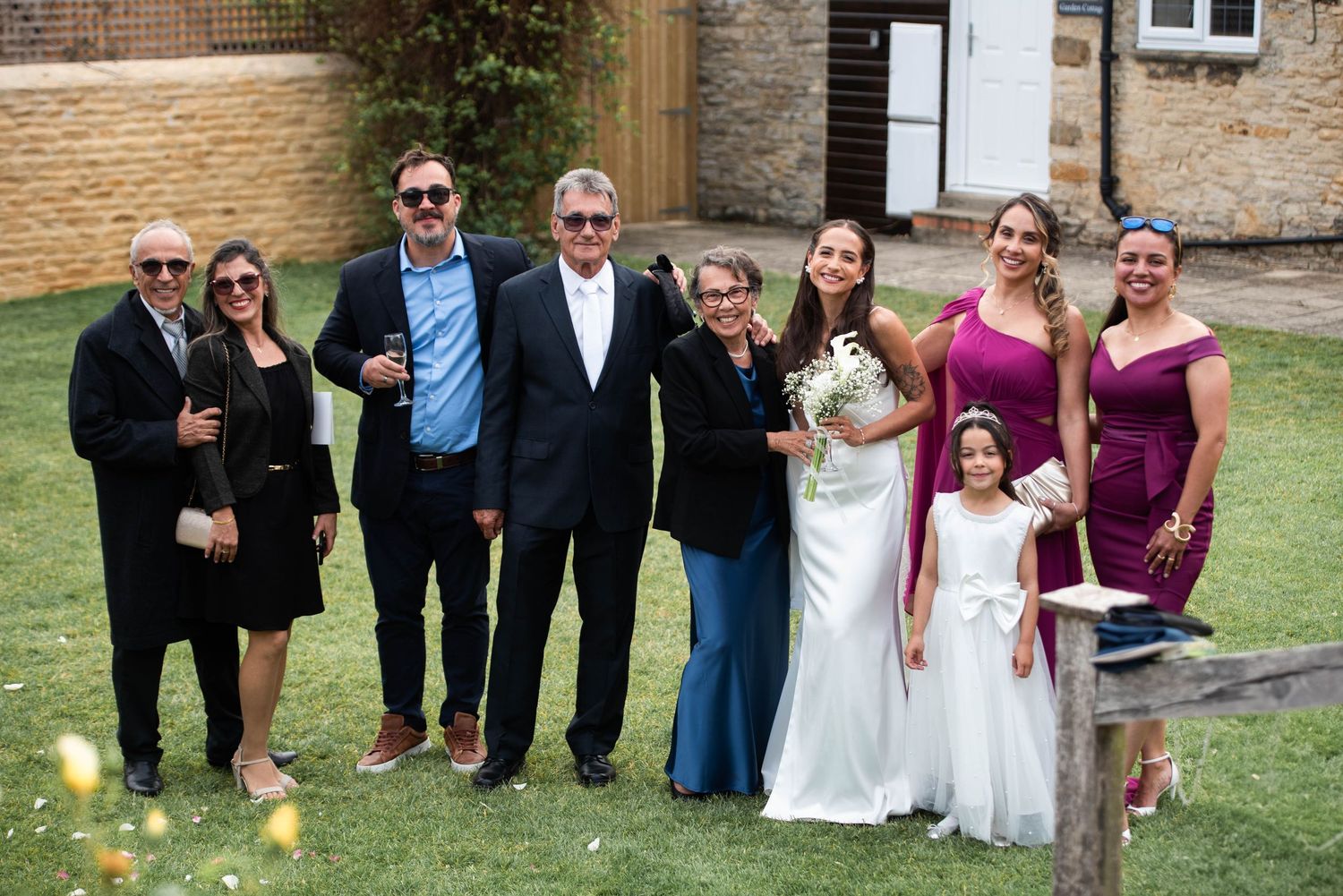 Wedding party group photo with bride in white gown, flower girl, and family members in formal attire outdoors.