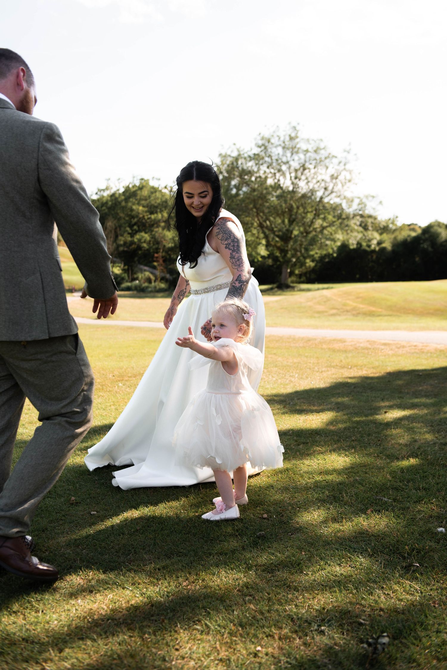 Bride in white gown with tattoos walks with toddler flower girl in pink tutu dress on green lawn.