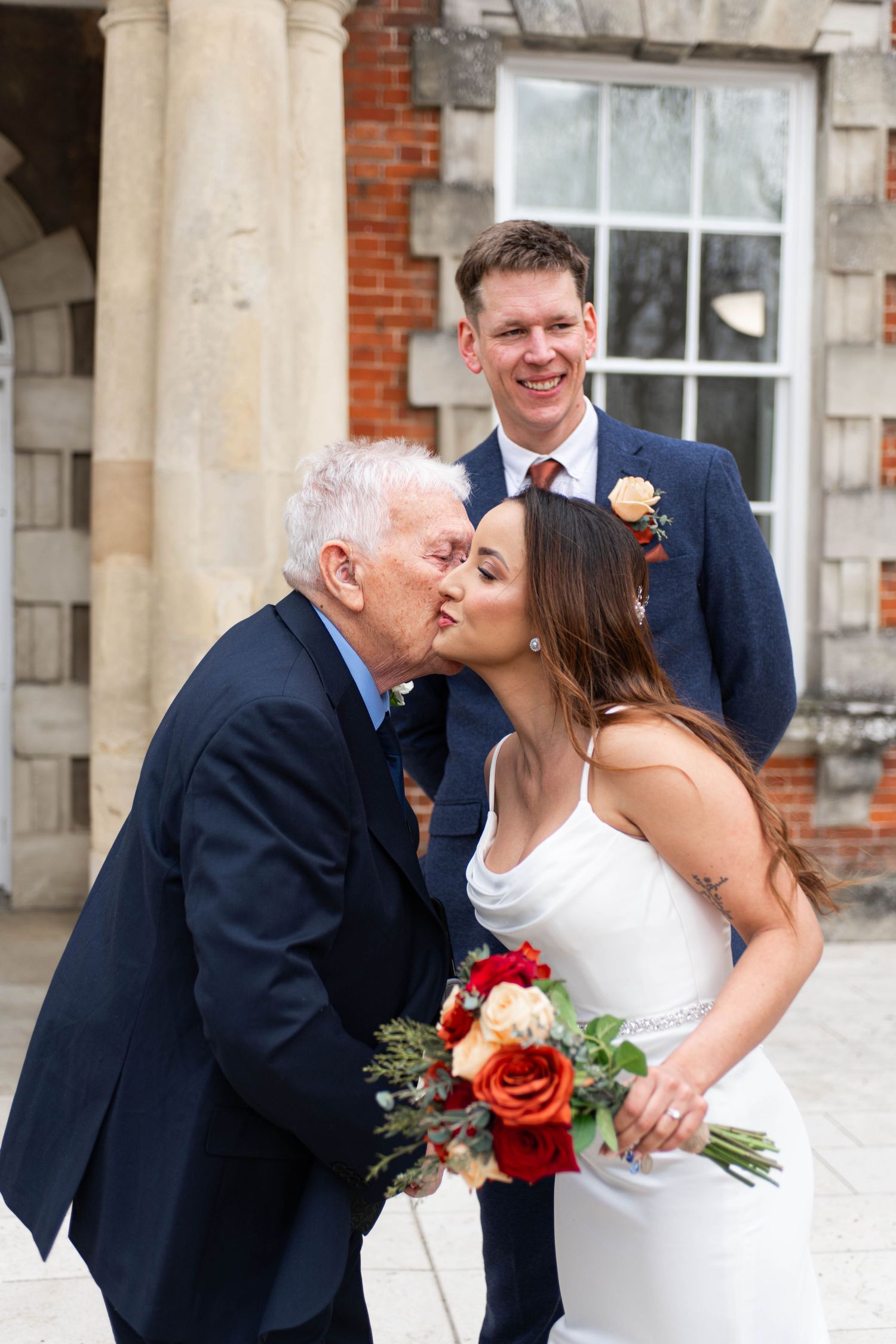 Elderly guest kisses bride on cheek as smiling groom looks on outside historic brick building.