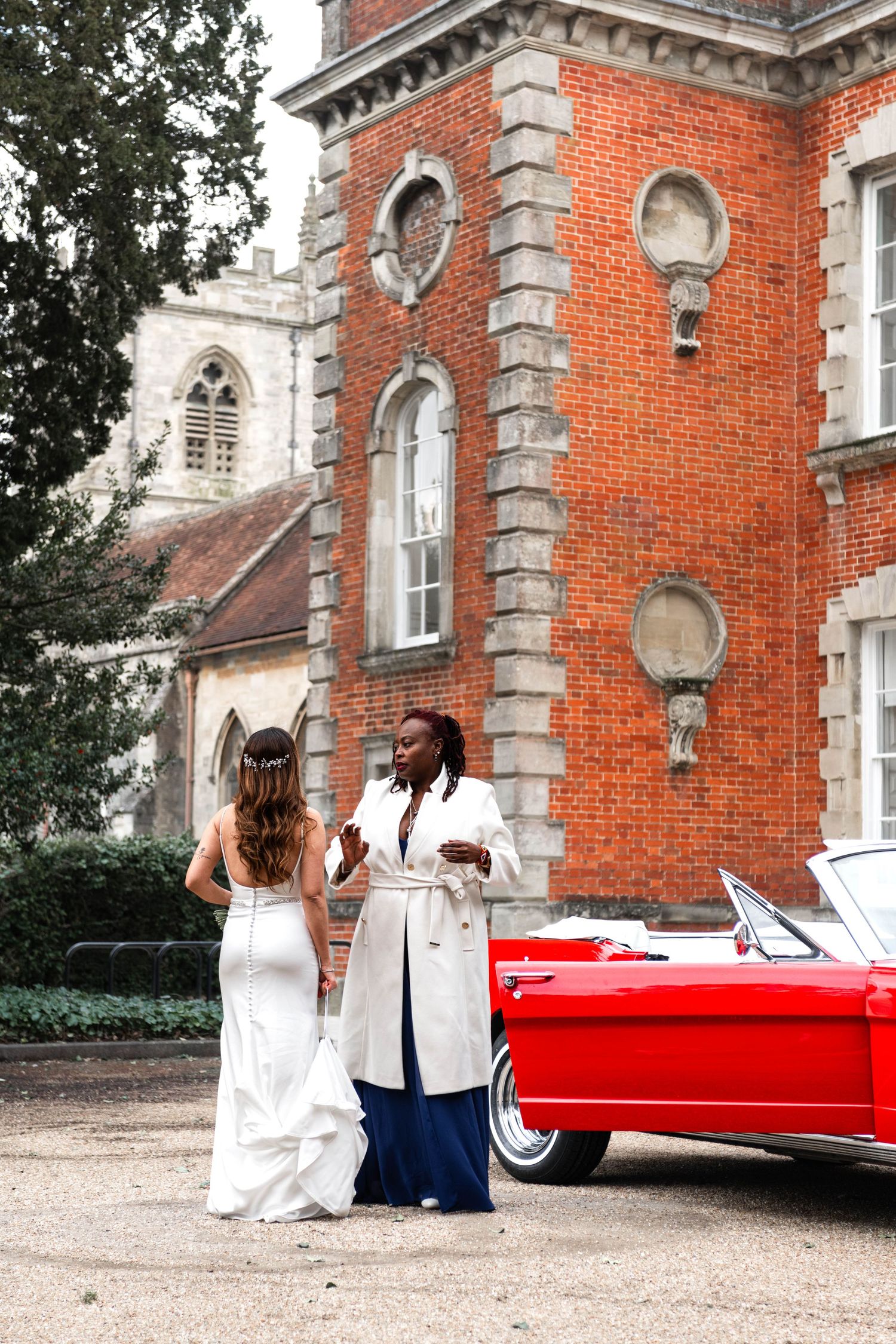 Bride in white gown chats with elegant guest in white coat beside red convertible at venue.