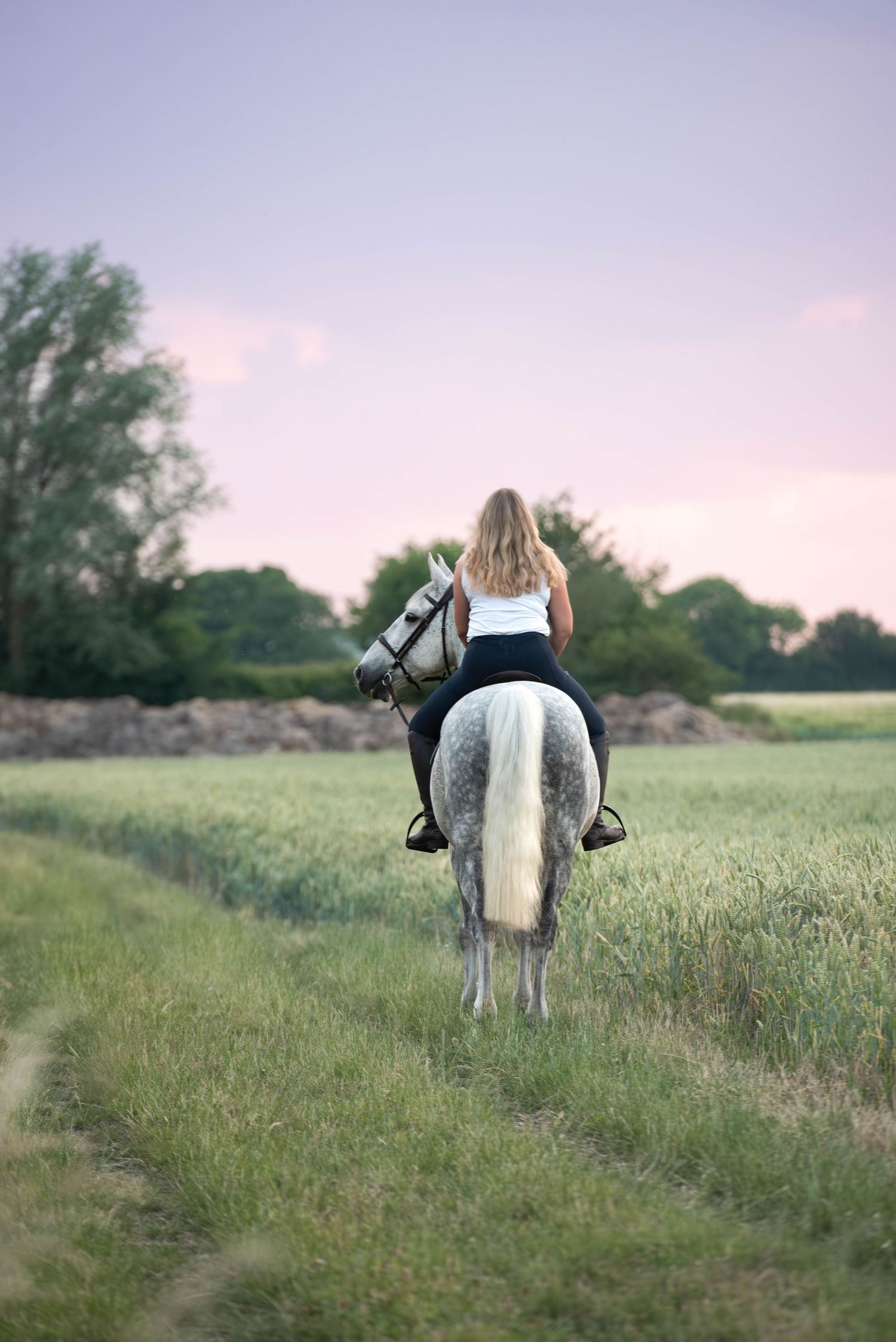 Blonde woman riding grey horse through green field at dusk from behind.