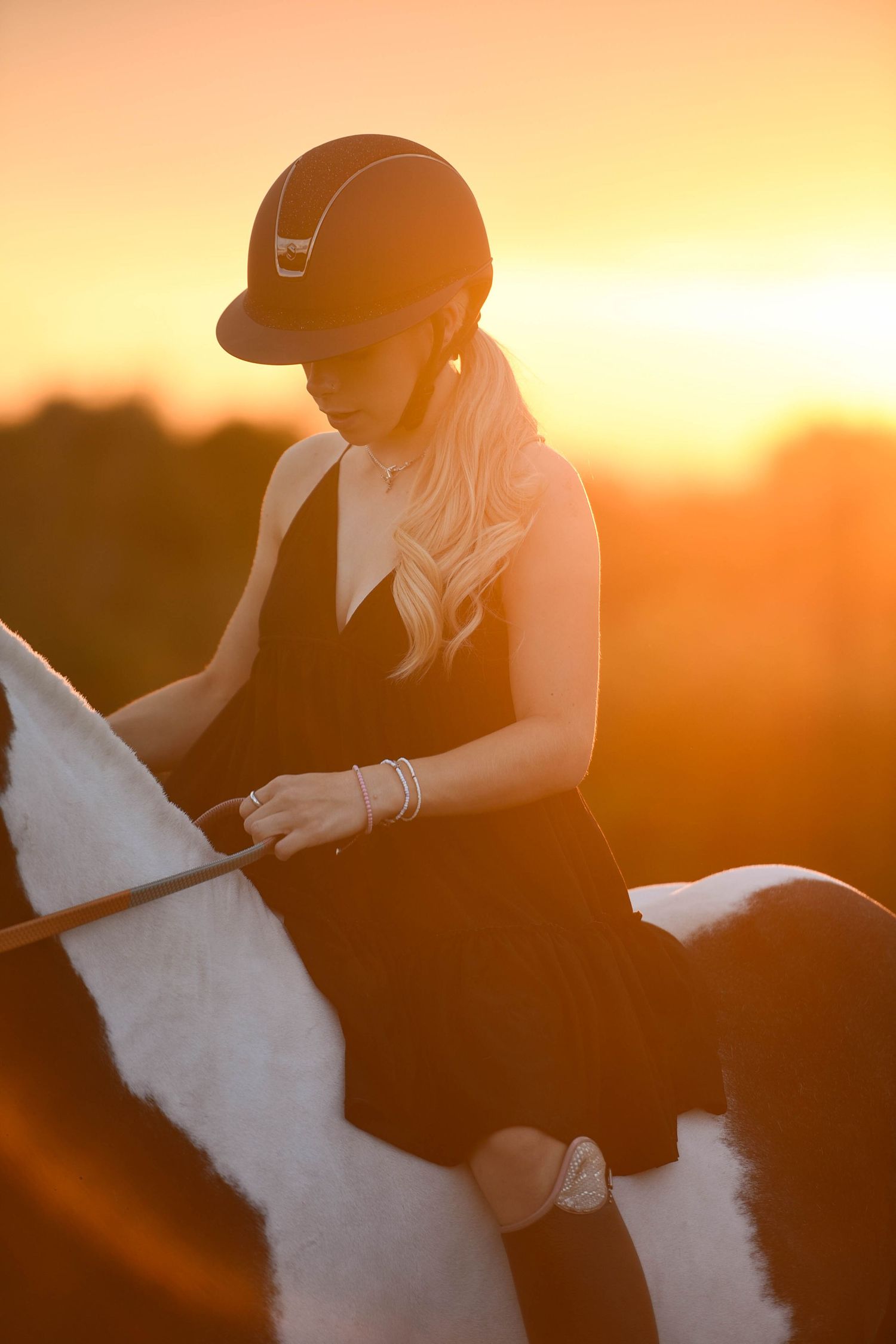 Blonde equestrian in black dress and riding helmet atop a paint horse during a vibrant golden sunset.