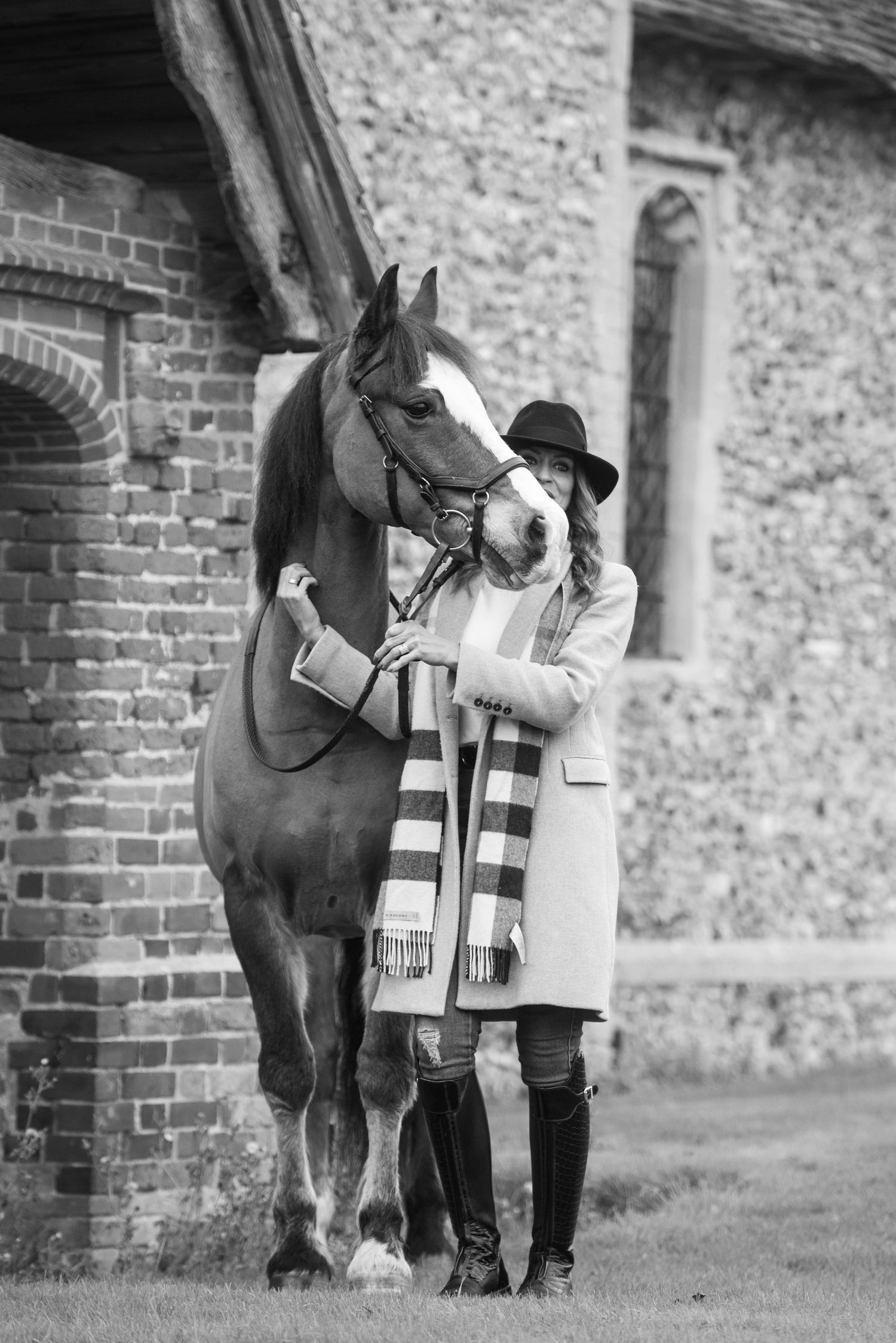 Black and white photo of a woman in a hat and scarf standing with a horse near a brick building.