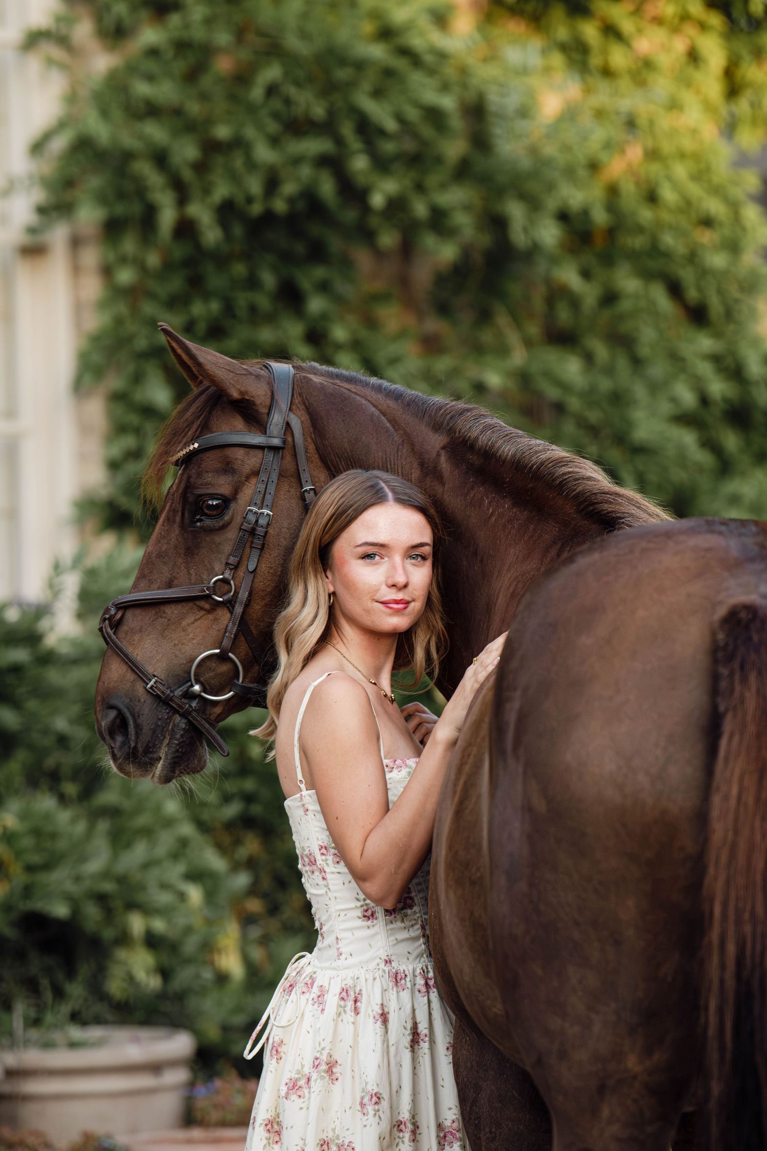 Young woman in floral dress standing beside a dark brown horse with bridle outdoors near green foliage.