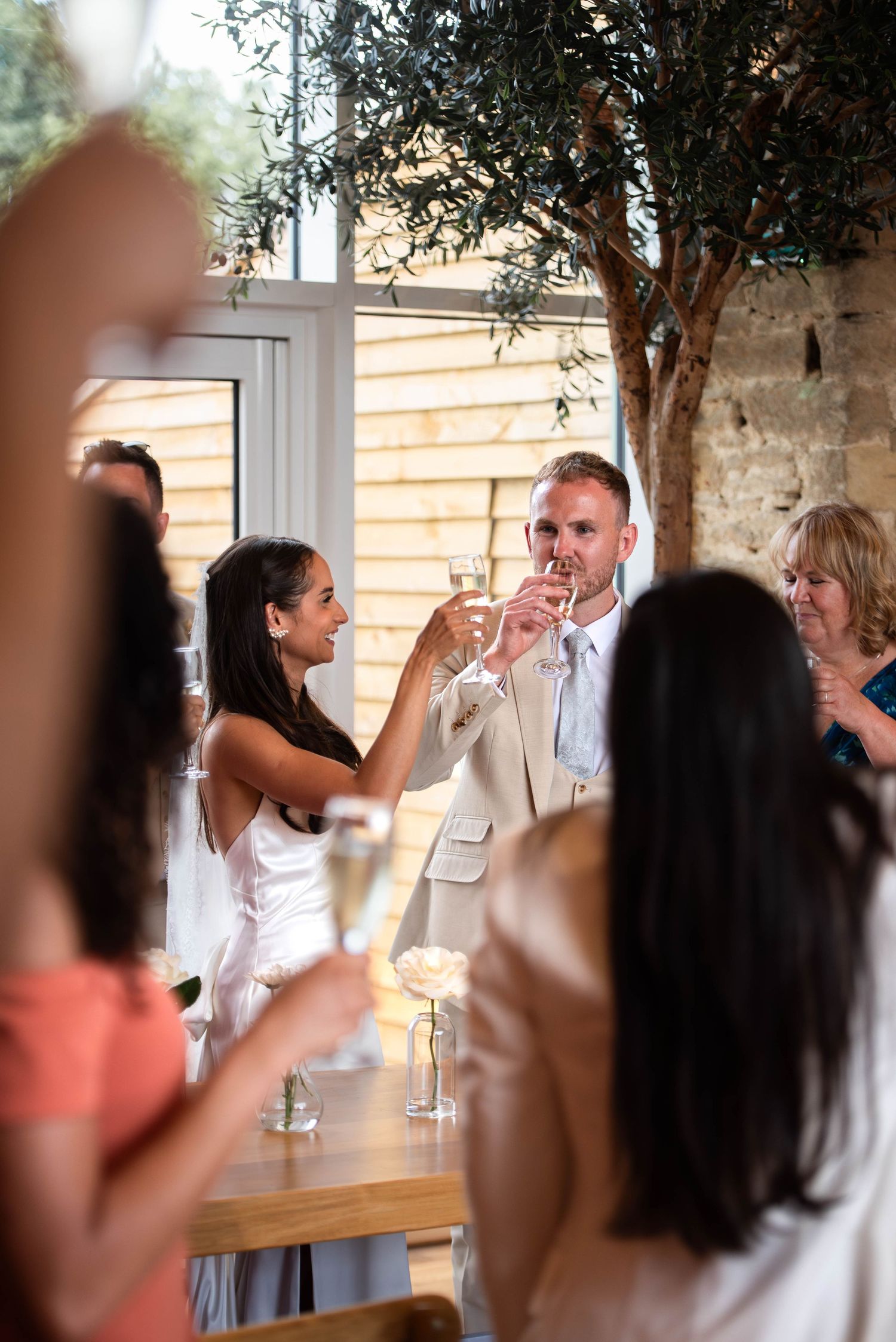 A bride and groom toast with champagne glasses surrounded by smiling wedding guests indoors.