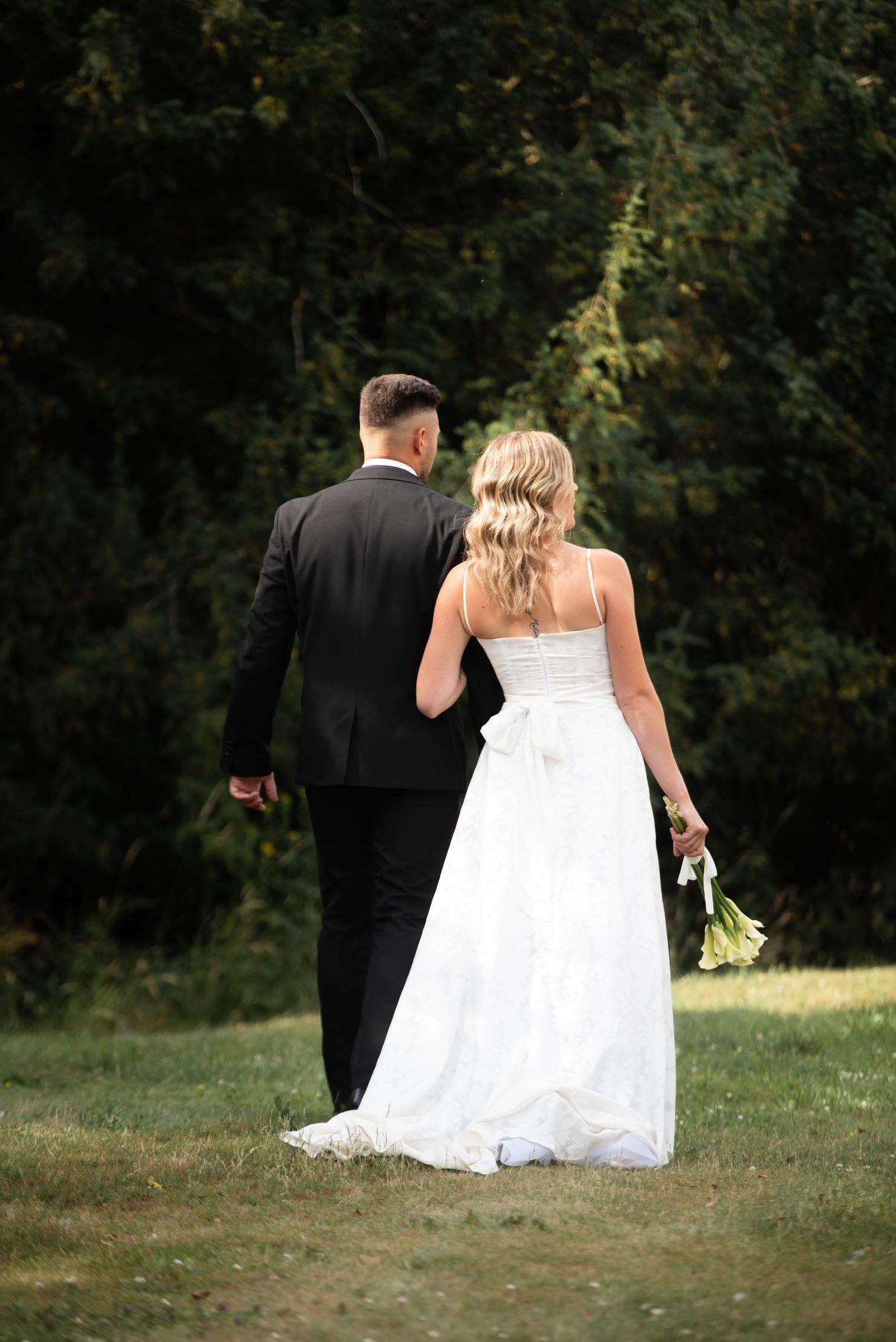 Bride in strapless white gown and groom in black suit walk hand in hand toward lush green forest.