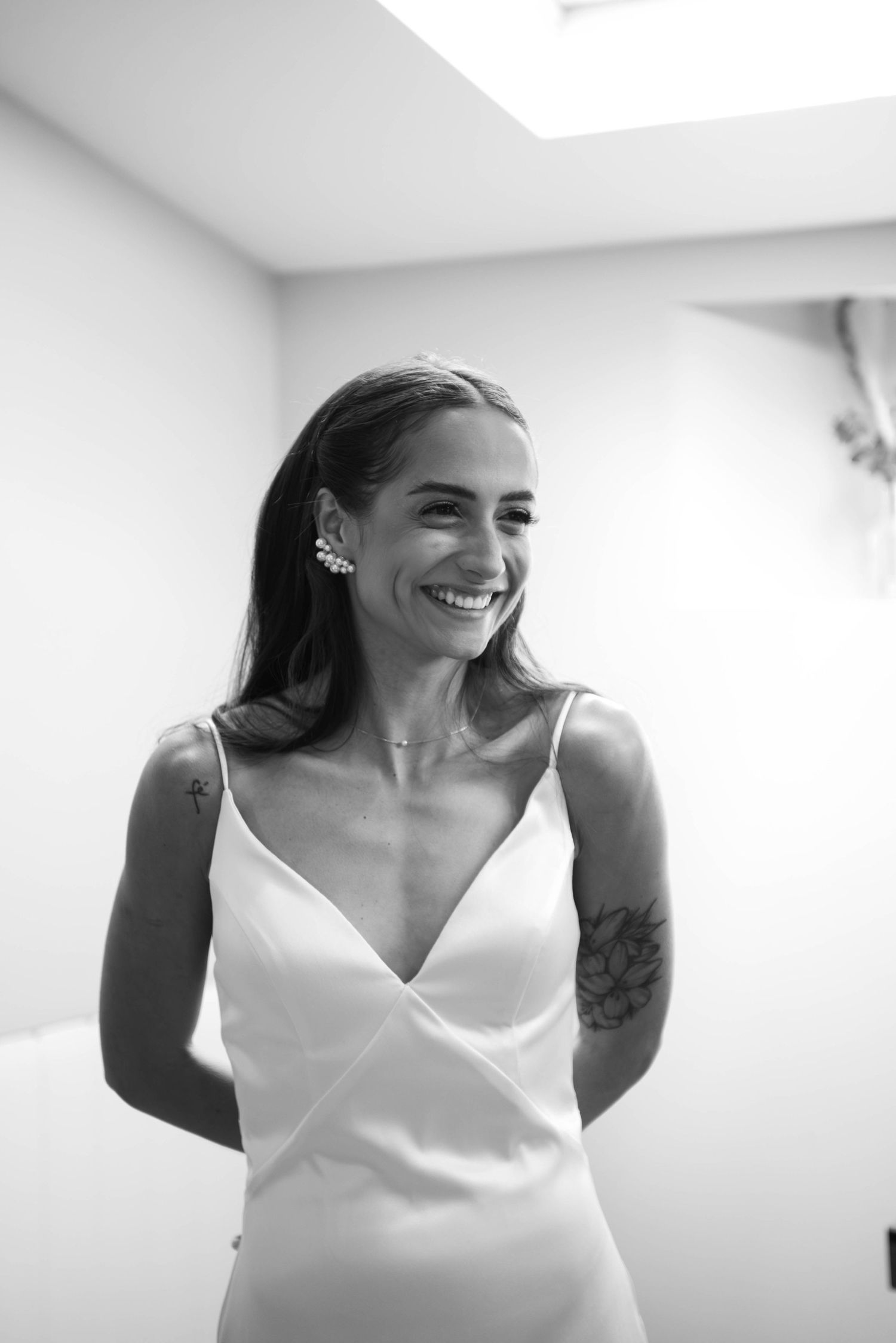 Black and white portrait of a smiling bride in a satin slip dress with floral tattoo and pearl earrings.
