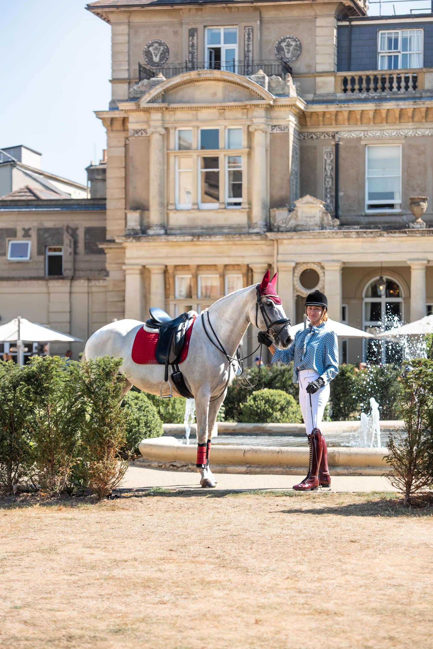 Equestrian rider in blue jacket standing beside a grey horse near a fountain outside a stately manor.