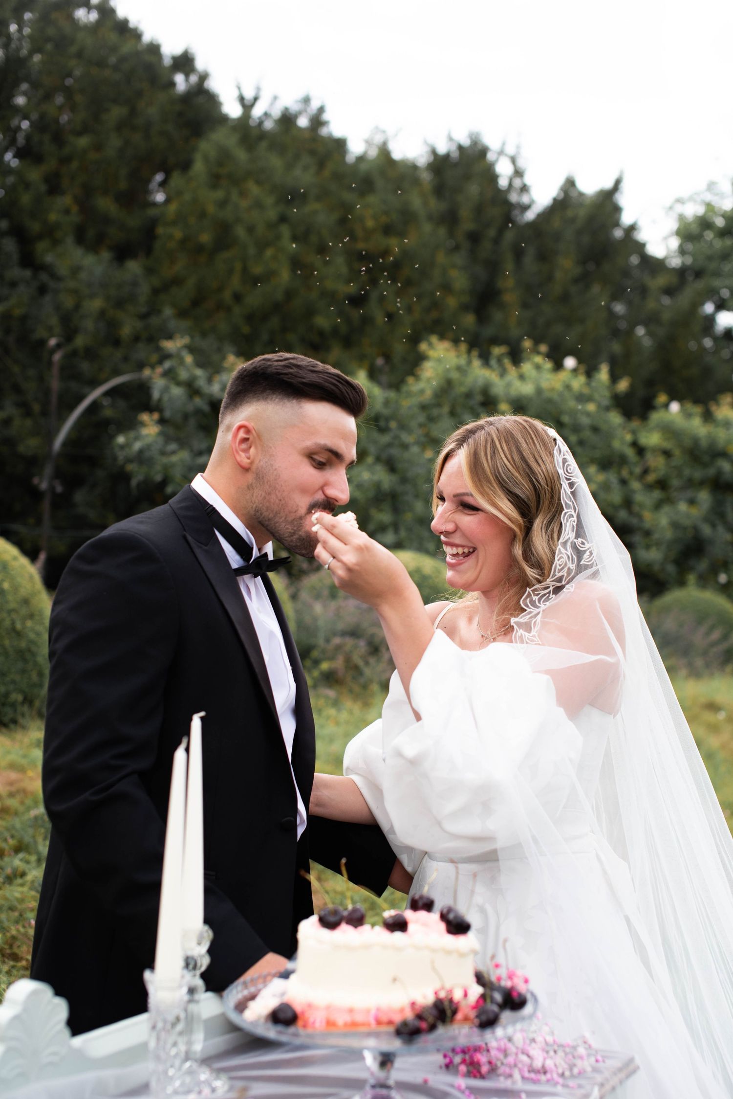 Smiling bride in puff-sleeve gown feeds wedding cake to groom in black tuxedo at outdoor reception.