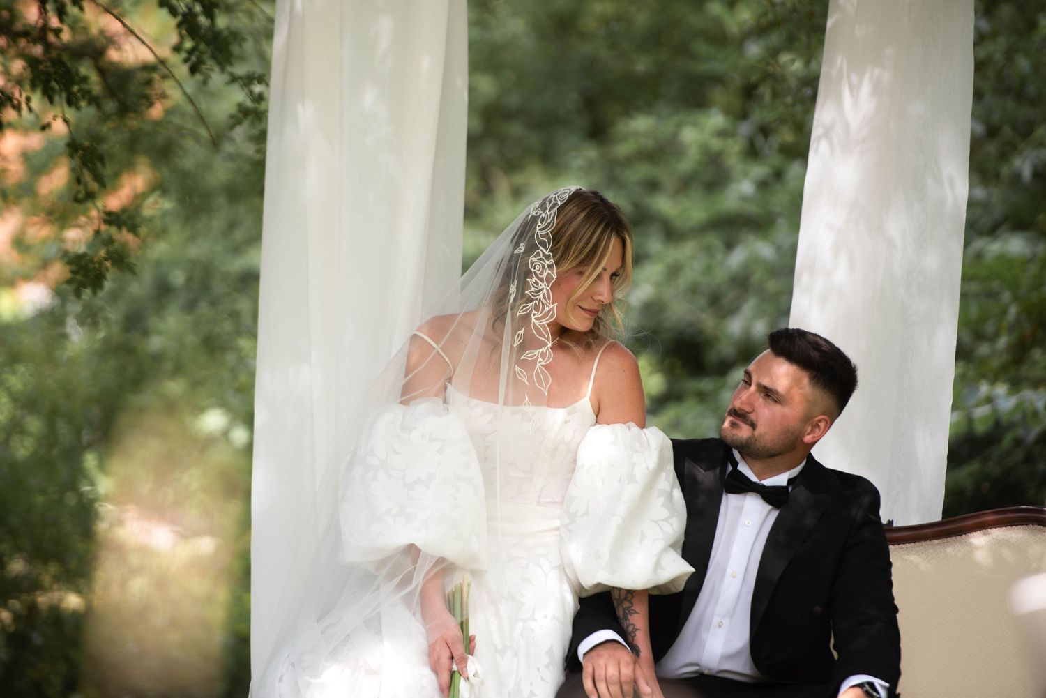 Bride and groom sharing intimate moment seated on ornate sofa under white draped outdoor wedding canopy.
