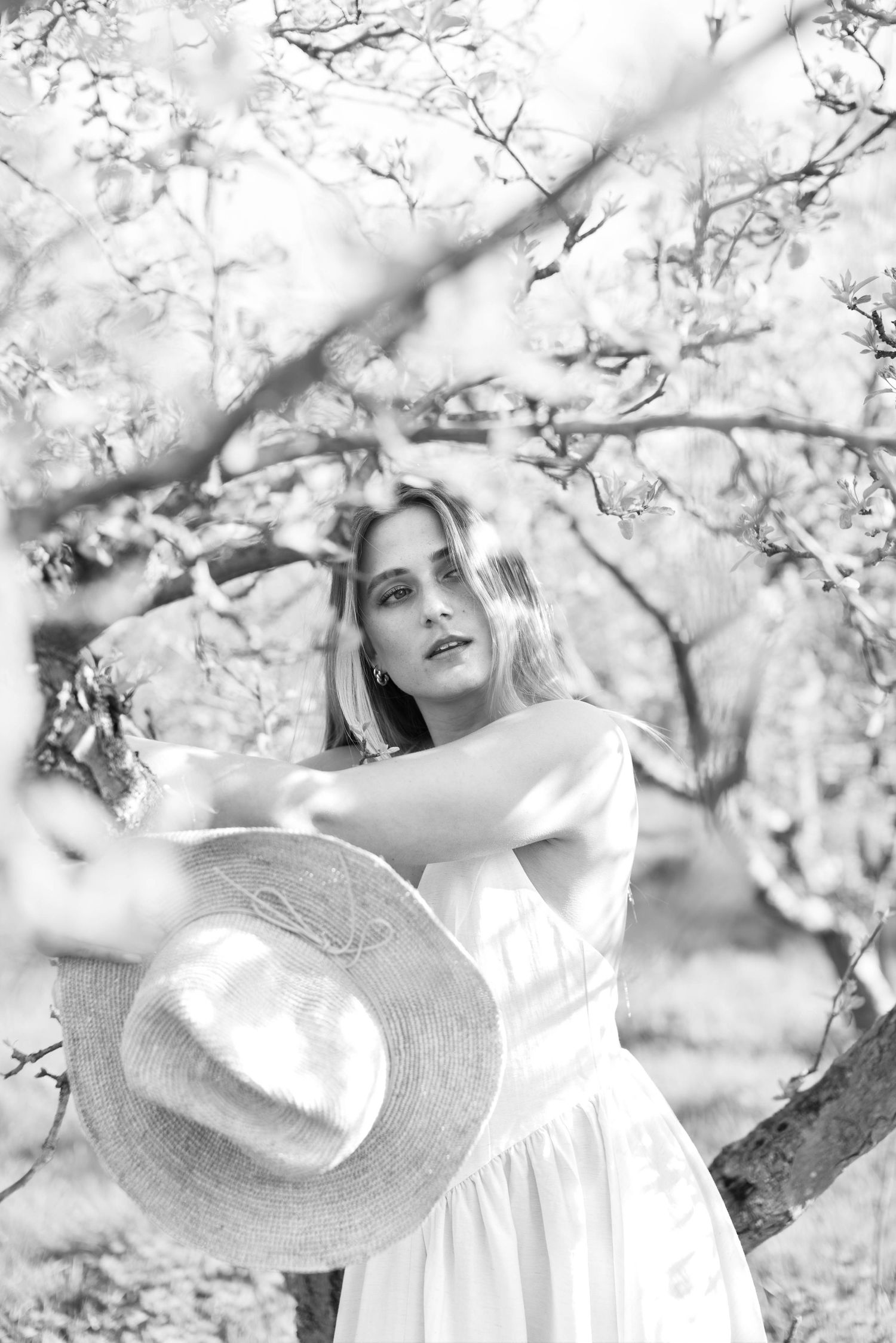 Black and white portrait of a blonde woman in white dress holding a hat among blossoming trees.