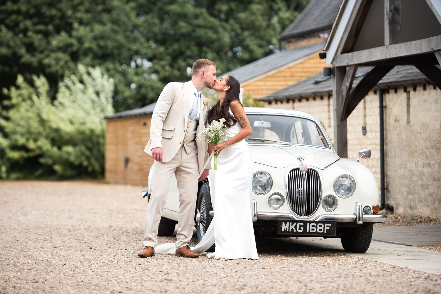 Bride and groom kissing beside a classic white Jaguar  vintage car at a rustic barn wedding venue.