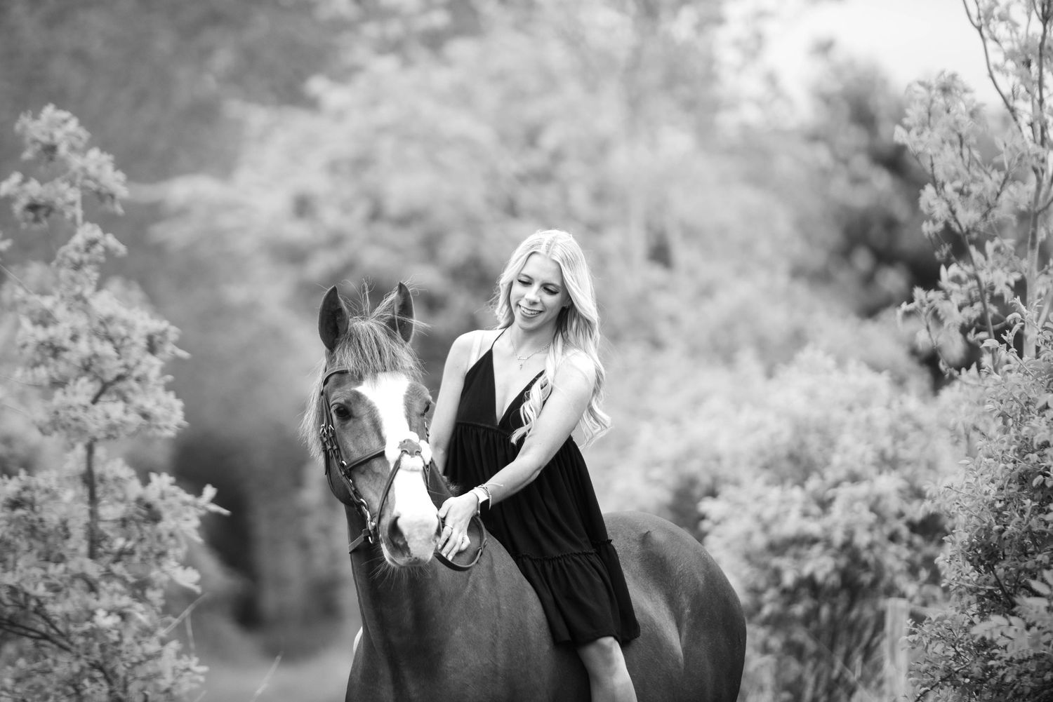 Black and white photo of a blonde woman in a black dress posing with a chestnut horse outdoors.