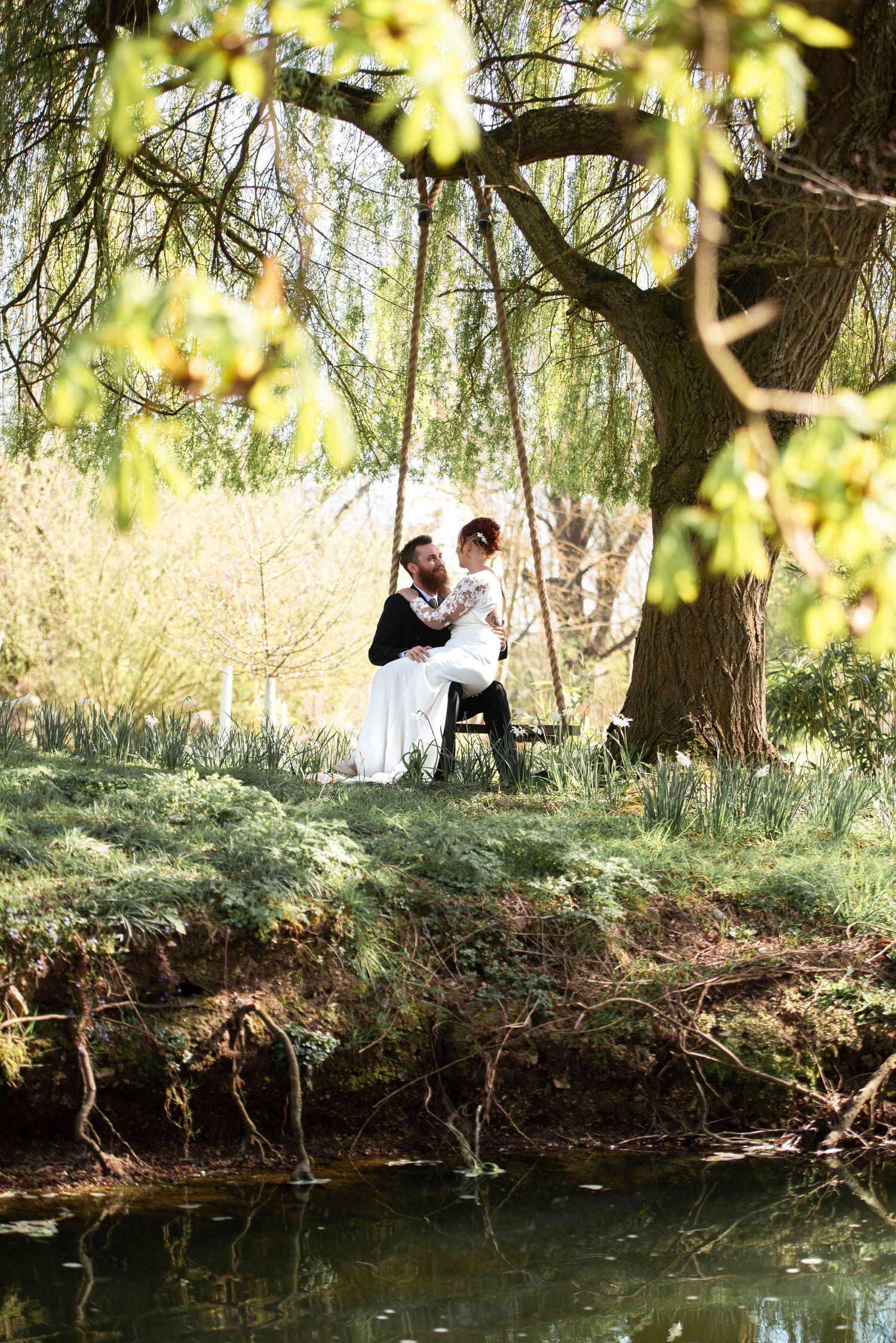 Romantic bride and groom embrace on a swing under a weeping willow tree beside a tranquil pond.