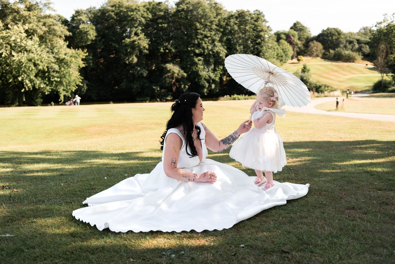 Bride in white gown sits on grass with toddler girl holding parasol umbrella in sunny park setting.