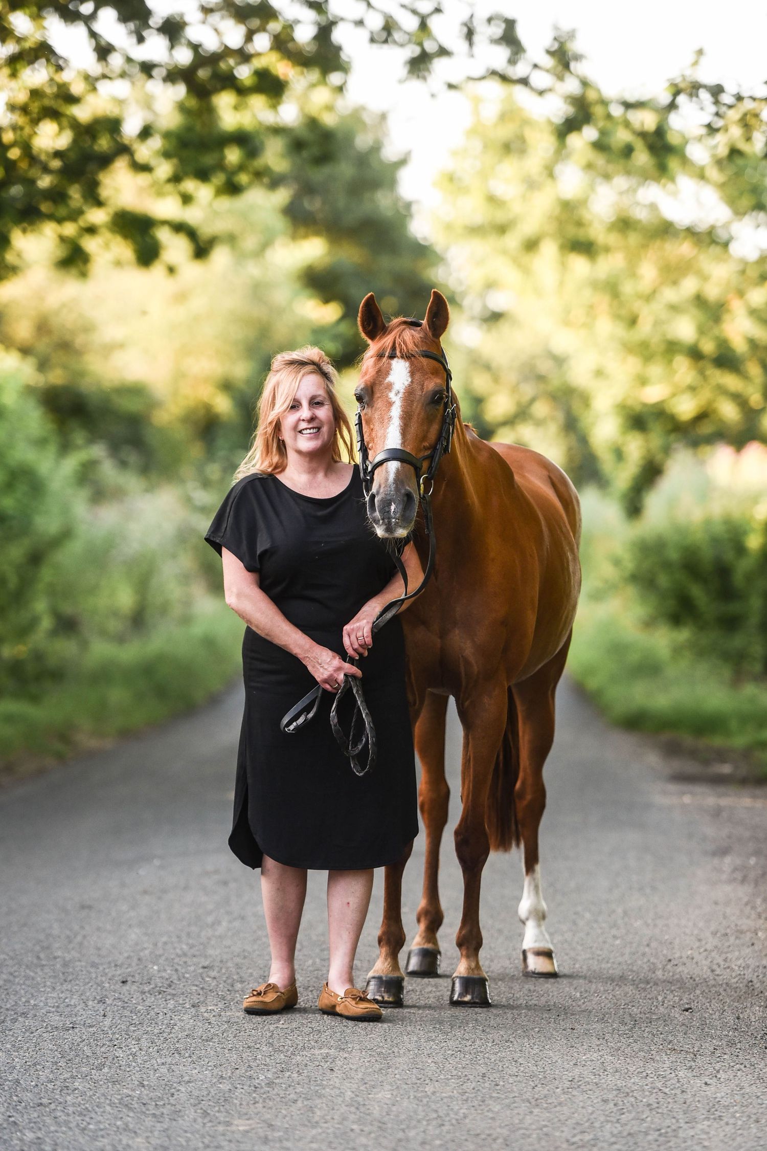 Smiling woman in black dress walking alongside a chestnut horse on a peaceful tree-lined rural road.