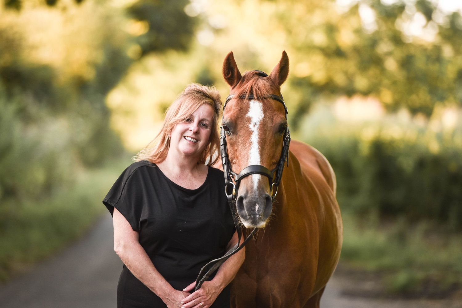 Blonde woman smiling closely beside a chestnut horse with a white blaze on a tree-lined road.