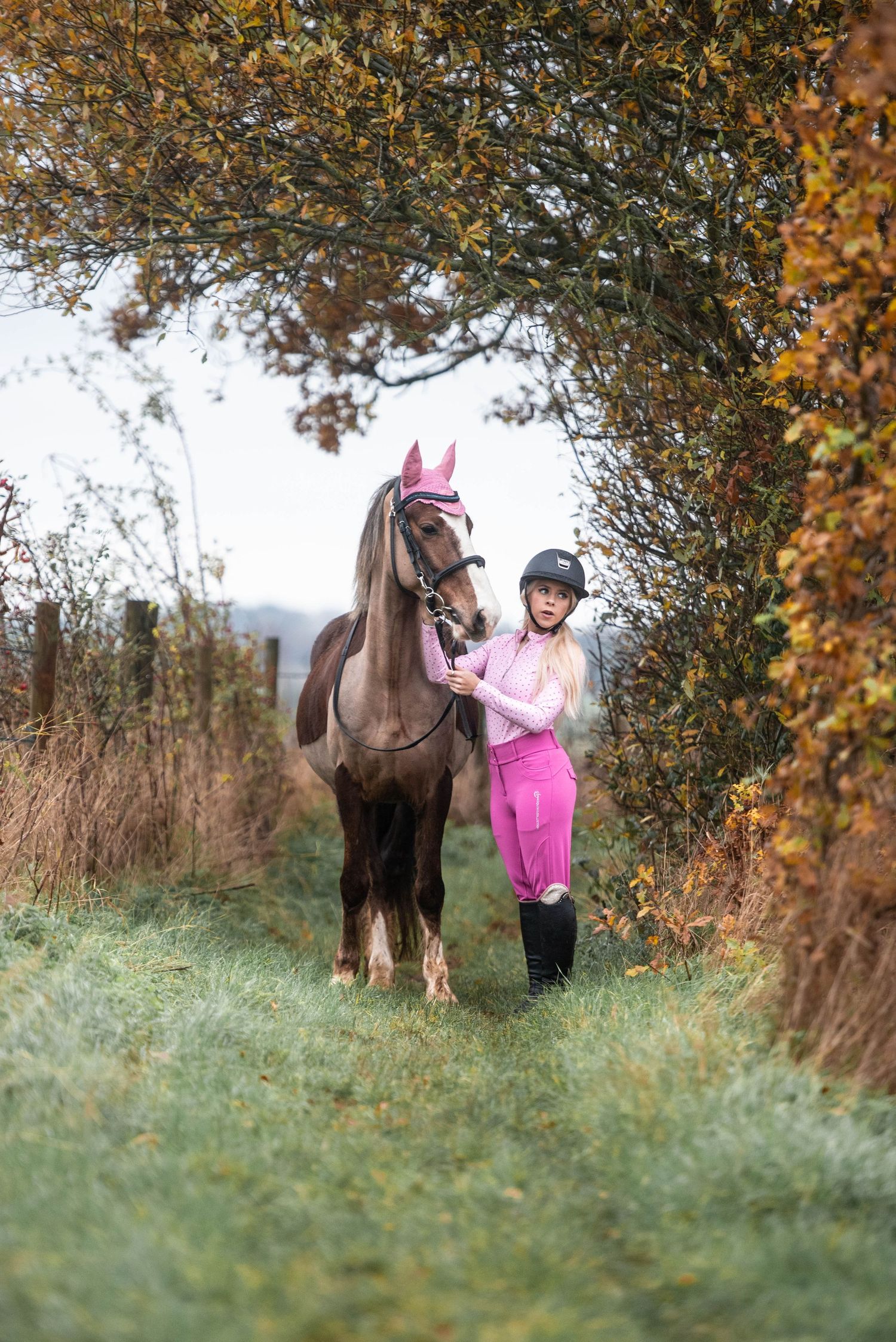 Blonde rider in pink outfit and helmet leading a brown horse through an autumn tree archway.