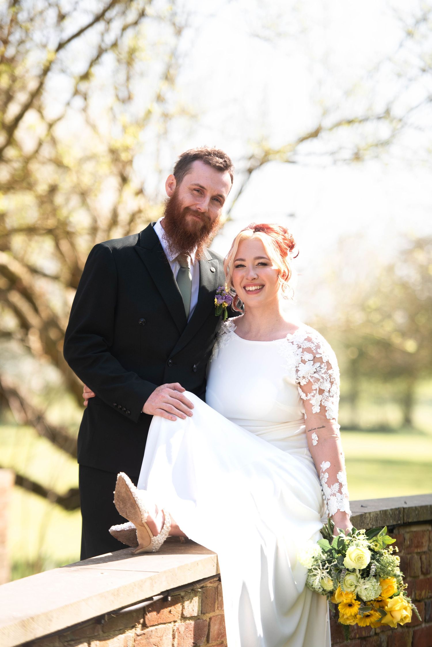 Smiling bride holding yellow sunflowers sits on brick wall beside bearded groom in dark suit outdoors.