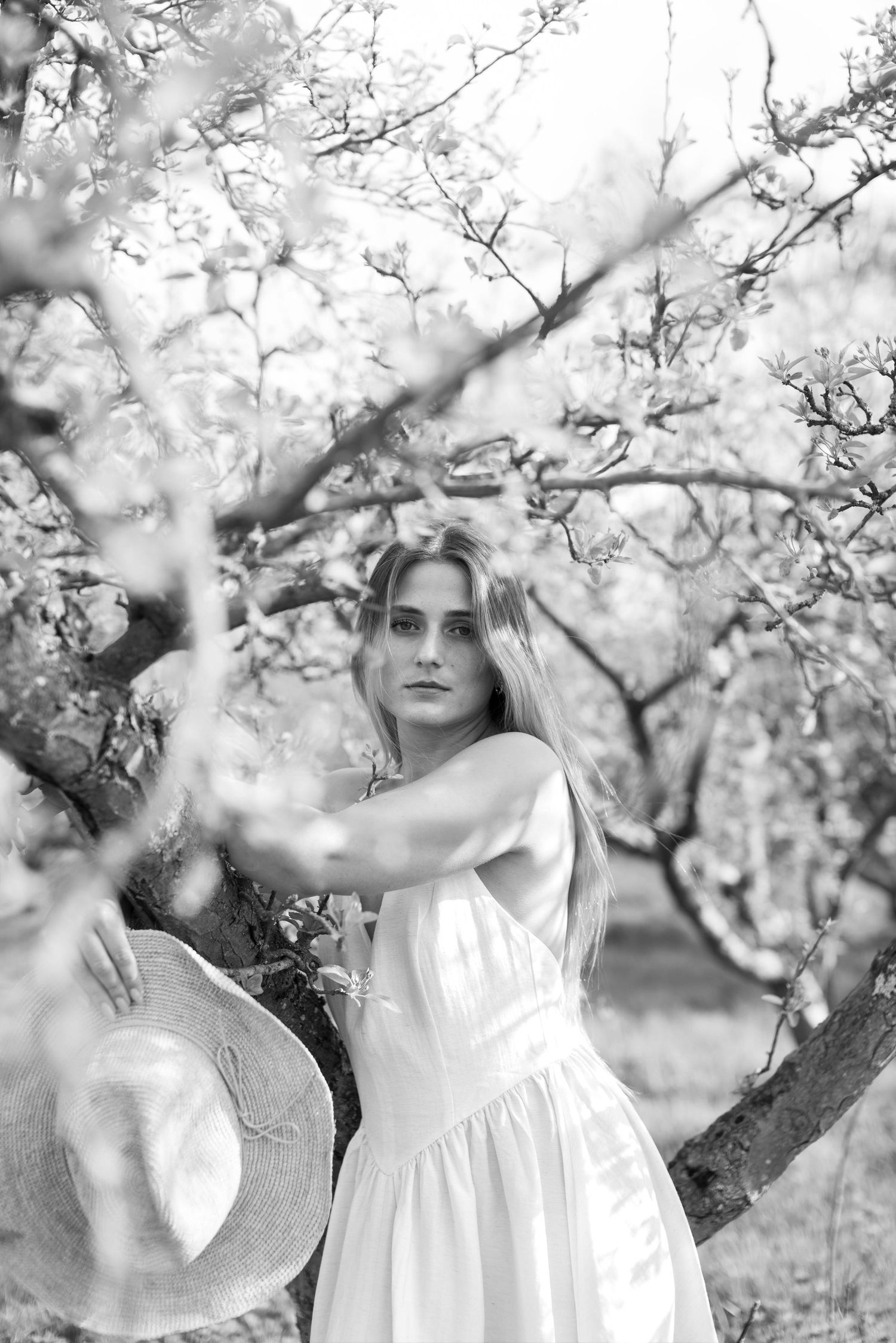 Blonde woman in white dress holds straw hat among flowering spring tree branches in black and white.