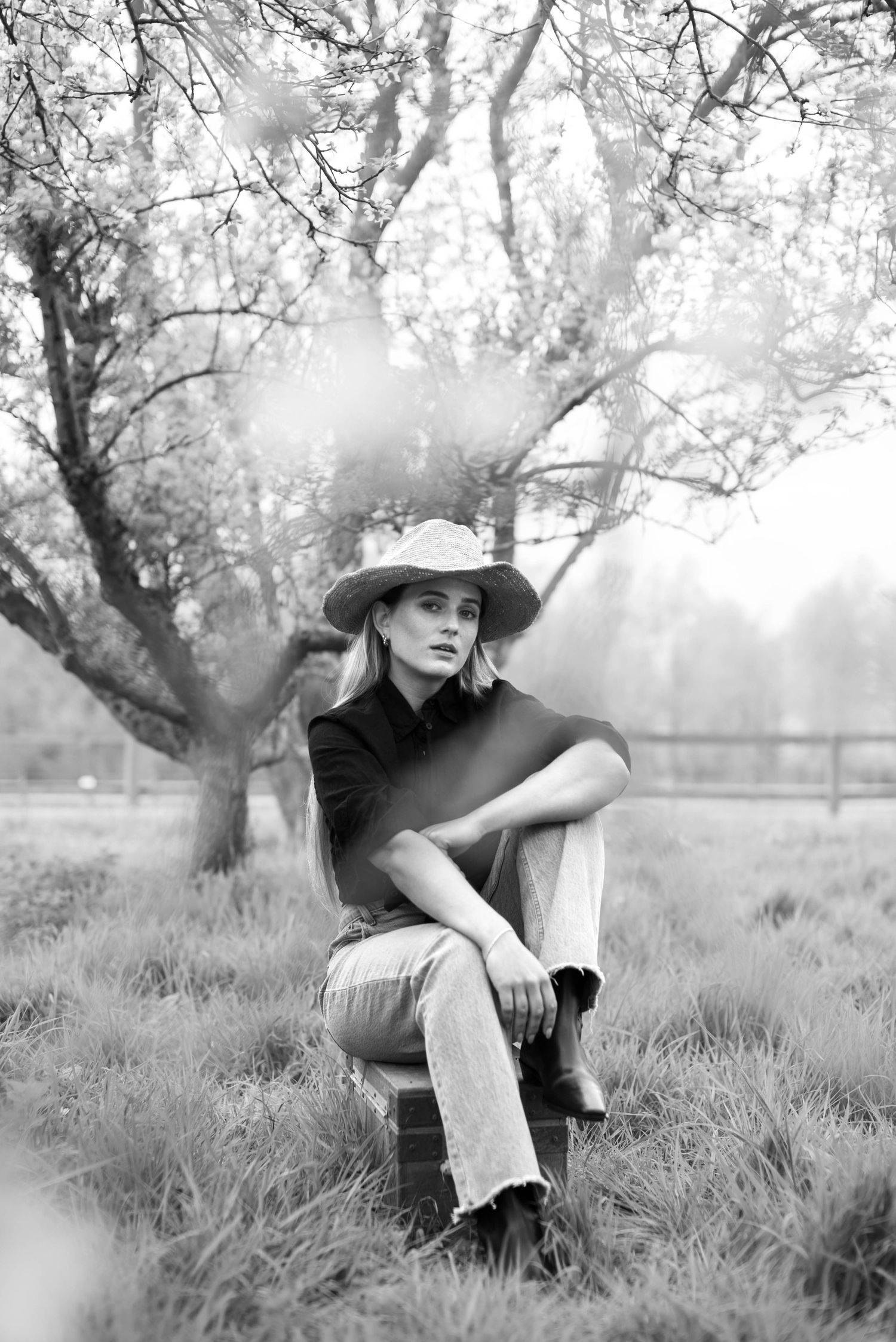 Black and white photo of woman in cowboy hat and black top sitting on chair in orchard field.