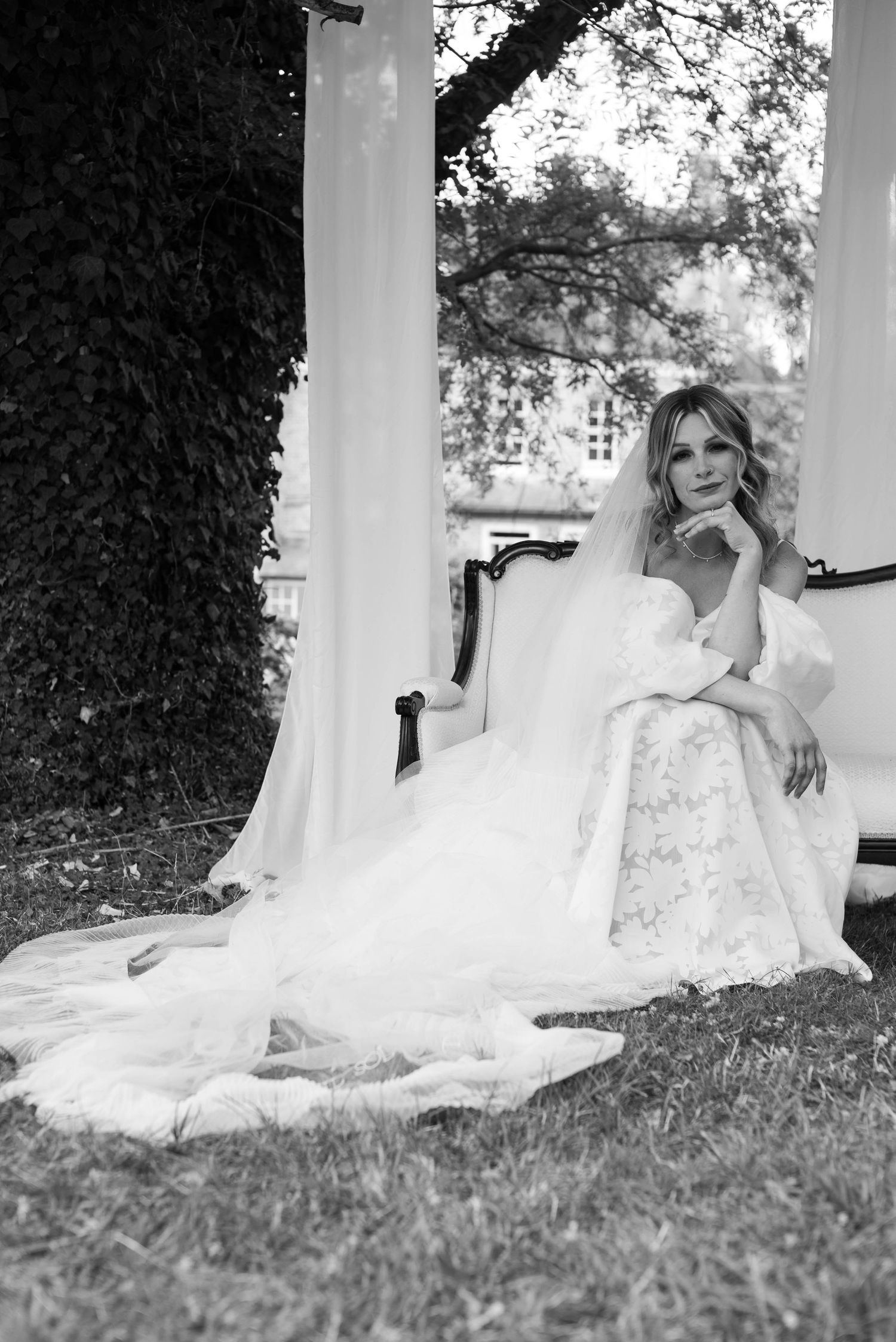 Elegant bride in lace wedding gown seated on vintage sofa under garden pergola, black and white portrait.