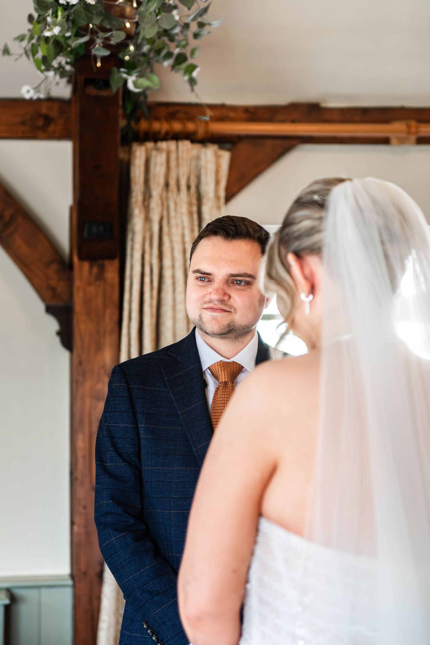 Groom in navy plaid suit with orange tie gazing lovingly at bride in white dress and veil during wedding ceremony.
