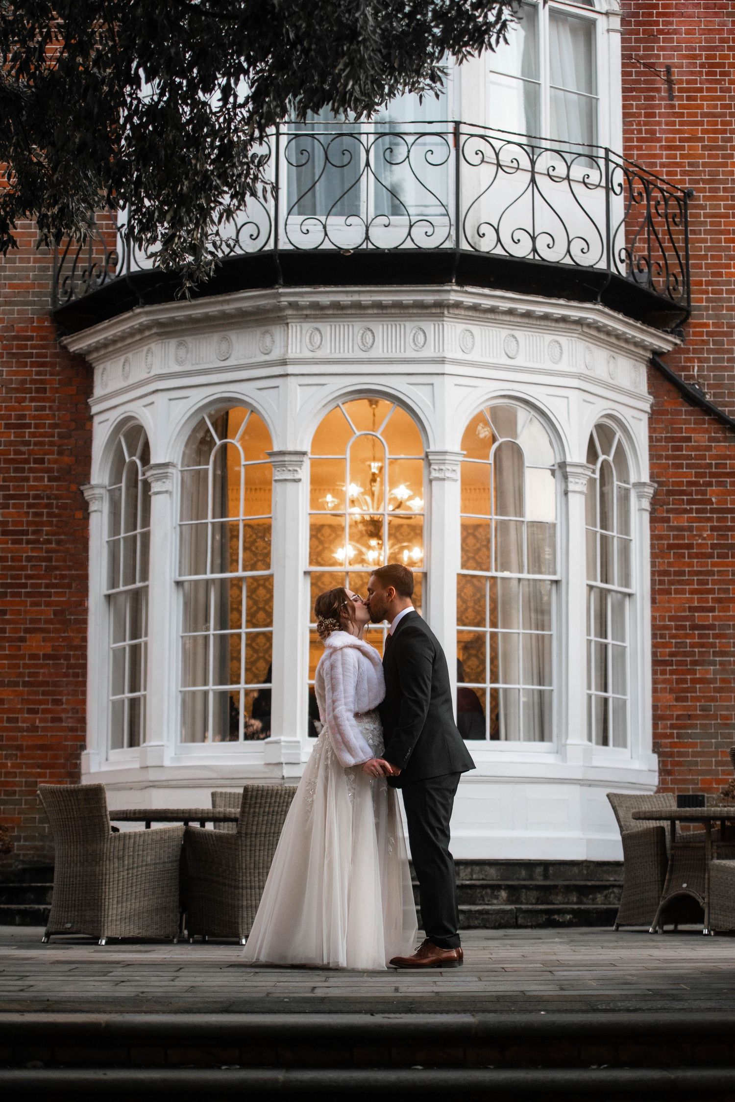 Bride and groom sharing romantic kiss outside elegant brick building with ornate bay window at dusk.